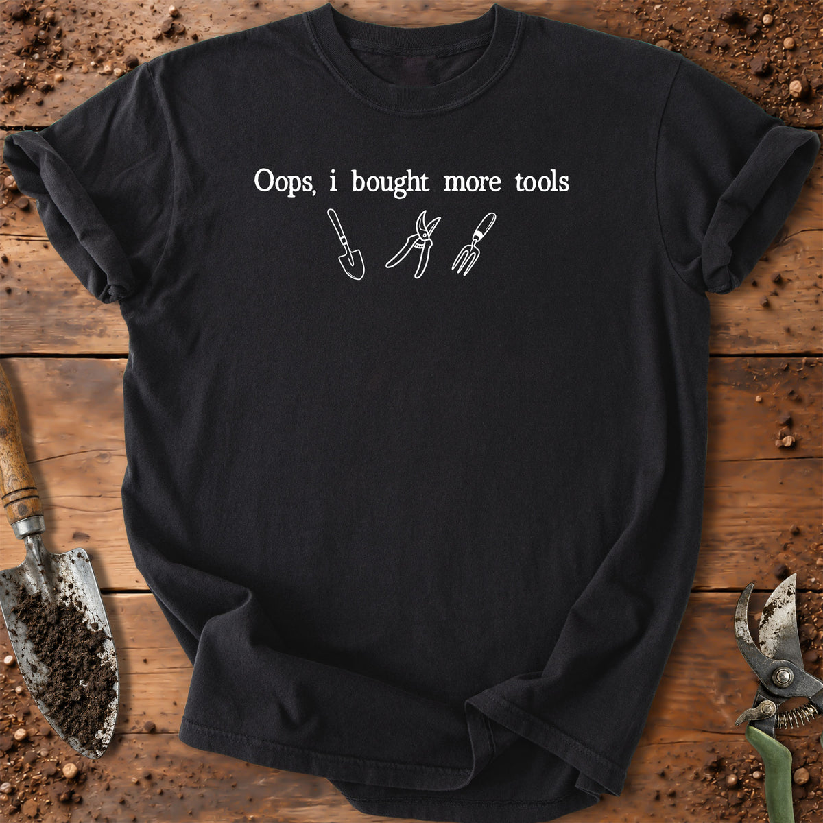 A black short-sleeve t-shirt with a white graphic print that reads 'Oops, i bought more tools' above simple line drawings of gardening tools. The shirt is laid flat on a rustic wooden surface surrounded by soil and gardening tools.