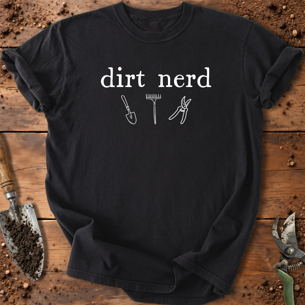 A black short-sleeve t-shirt with a white graphic that reads 'dirt nerd' above simple line drawings of a shovel, rake, and pruning shears, laid flat on a rustic wooden surface with soil and gardening tools around it.