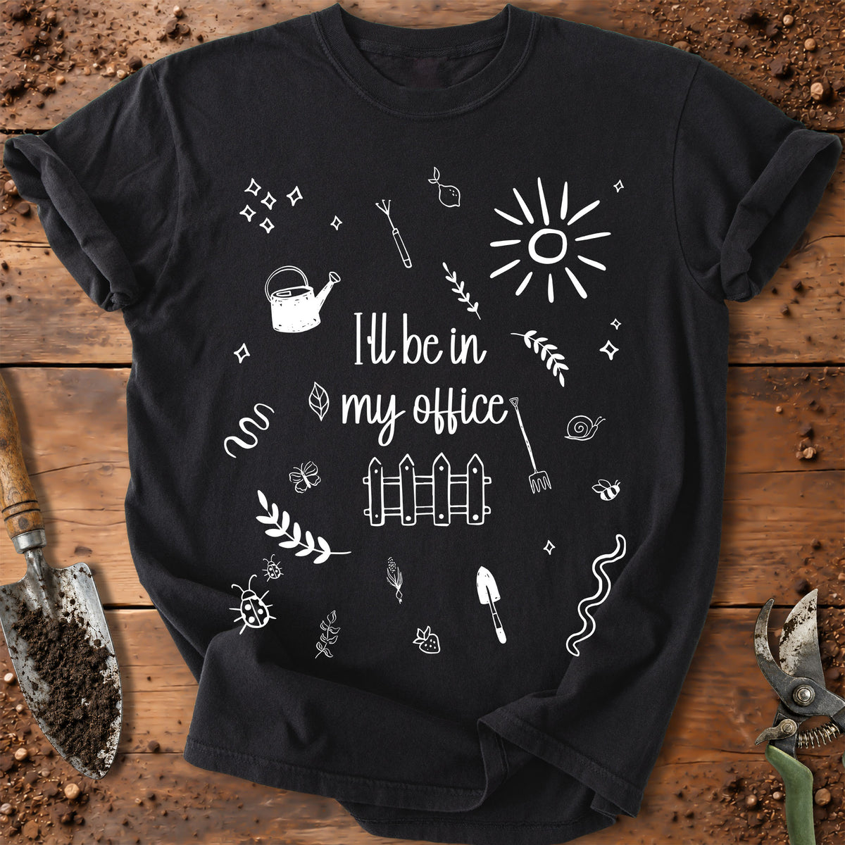 A black short-sleeve graphic T-shirt laid flat on a rustic wooden surface with gardening props like a trowel, soil, and pruning shears. The shirt has a white hand-drawn style print reading “I’ll be in my office” surrounded by doodles including a sun, watering can, fence, plants, and tools.