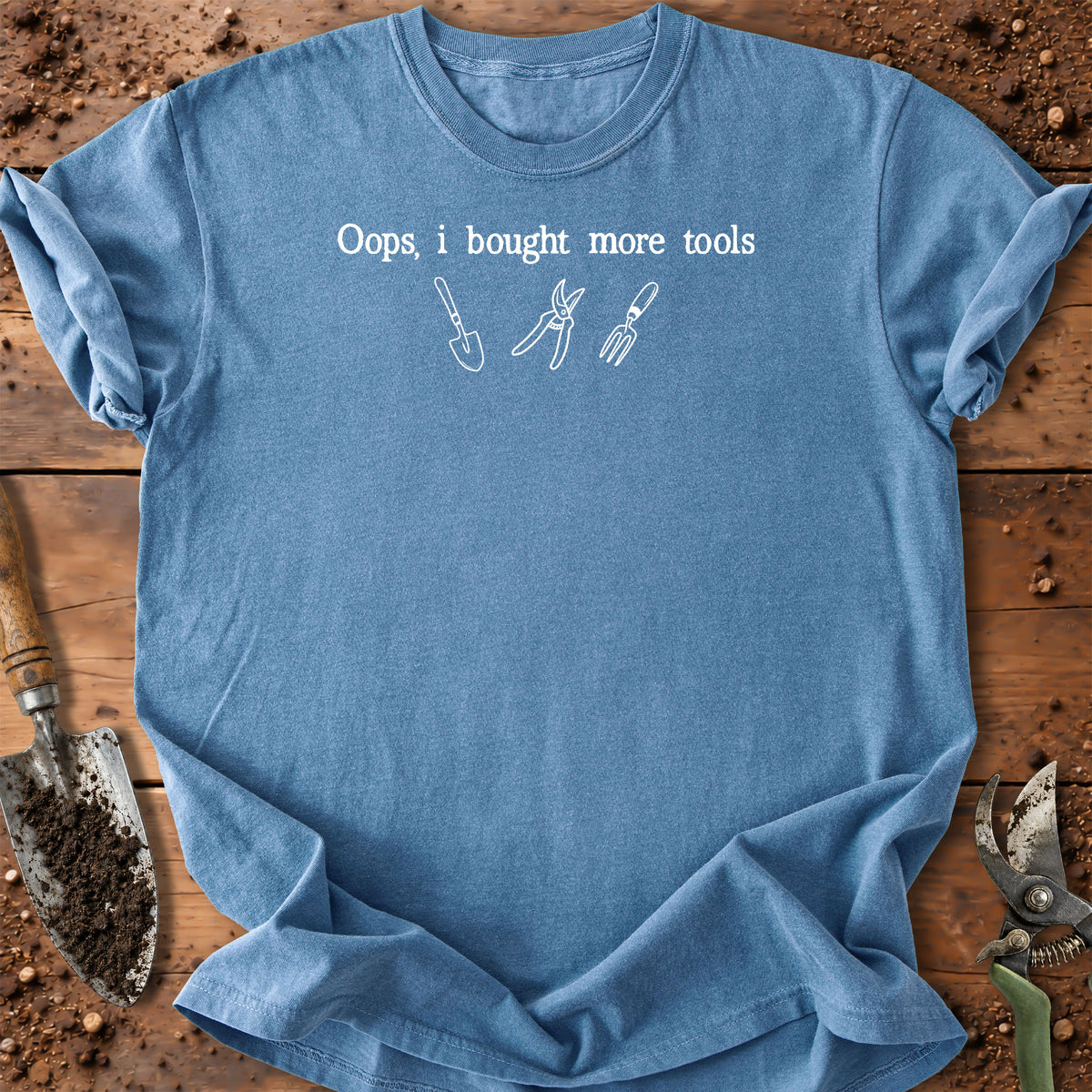 A light blue crewneck t-shirt with a white graphic print that reads 'Oops, i bought more tools' above simple line drawings of gardening tools. The shirt is laid flat on a rustic wooden surface surrounded by gardening props like soil, a trowel, and pruning shears.