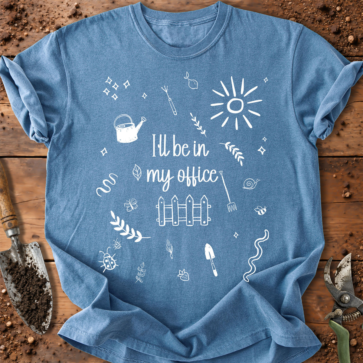 A blue short-sleeve crewneck t-shirt with a white graphic print that reads 'I'll be in my office' surrounded by gardening doodles like a watering can, fence, and sun. The shirt is laid flat on a rustic wooden surface with gardening tools like a trowel and pruning shears visible around the edges.