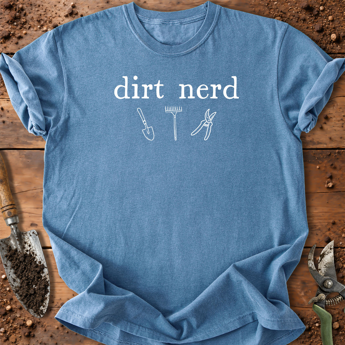 A light blue crewneck t-shirt with 'dirt nerd' printed in white text above gardening tool illustrations, laid flat on a rustic wooden surface with soil and tools around it.