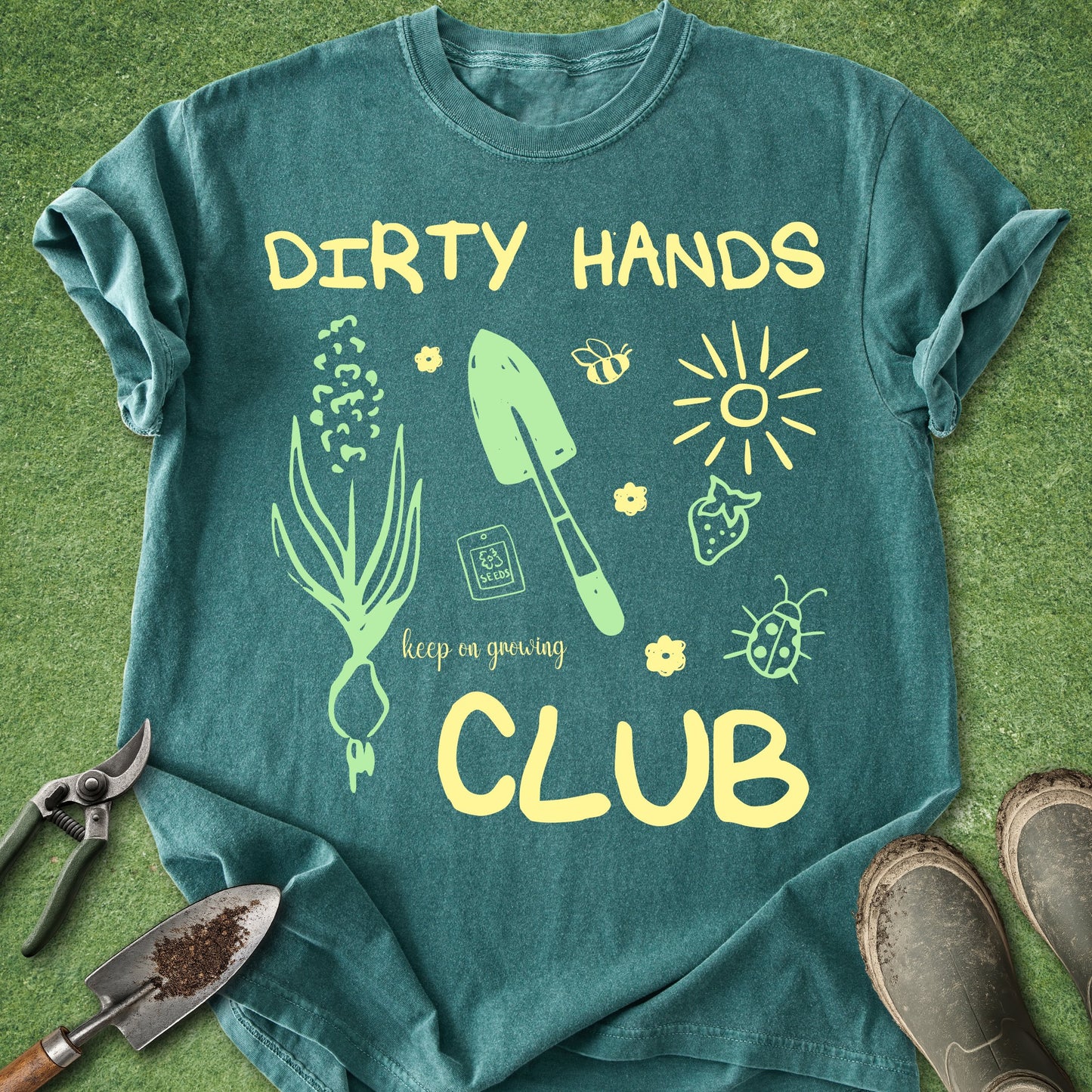Teal t-shirt with gardening-themed design and 'Dirty Hands Club' text on a grass background.