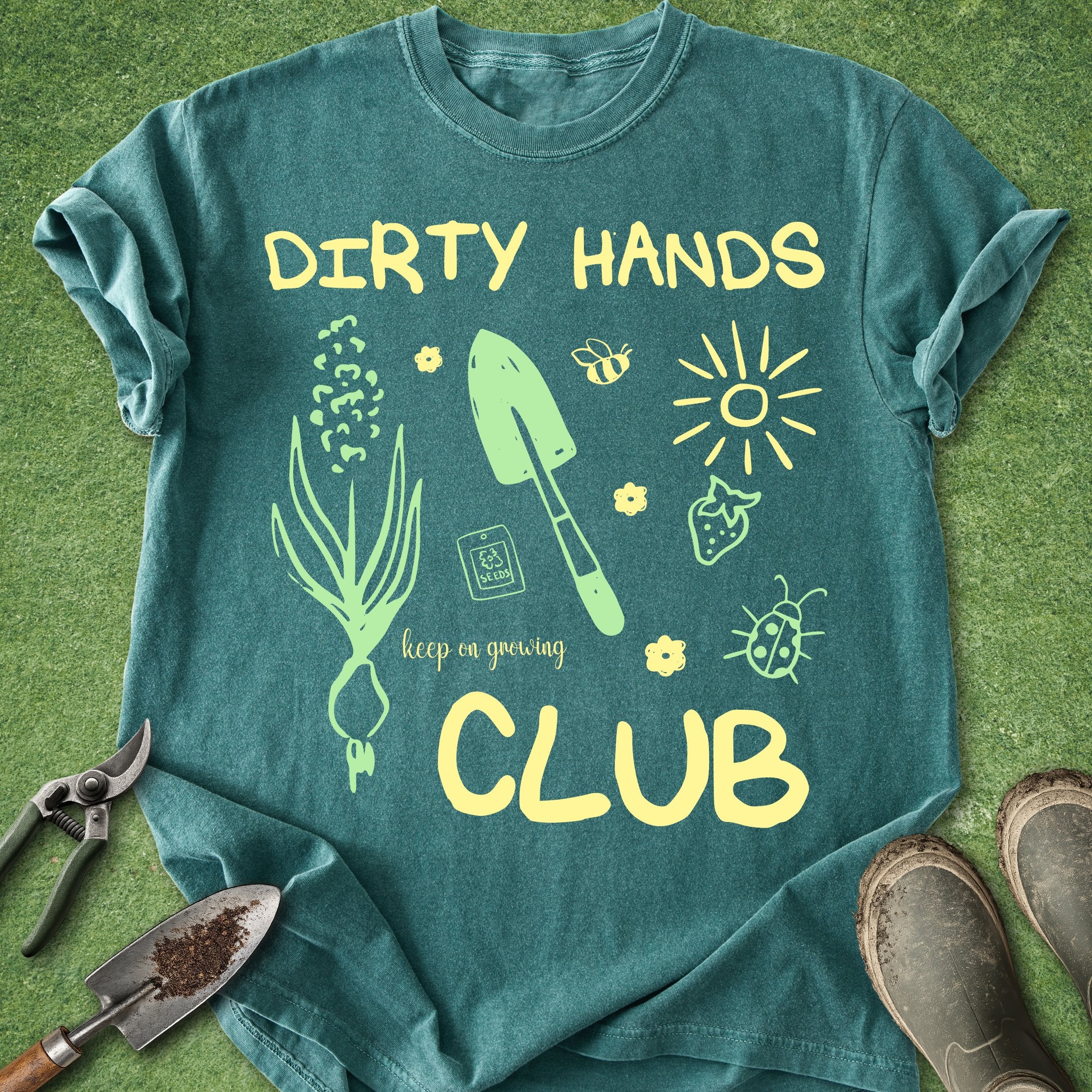 Teal t-shirt with gardening-themed design and 'Dirty Hands Club' text on a grass background.