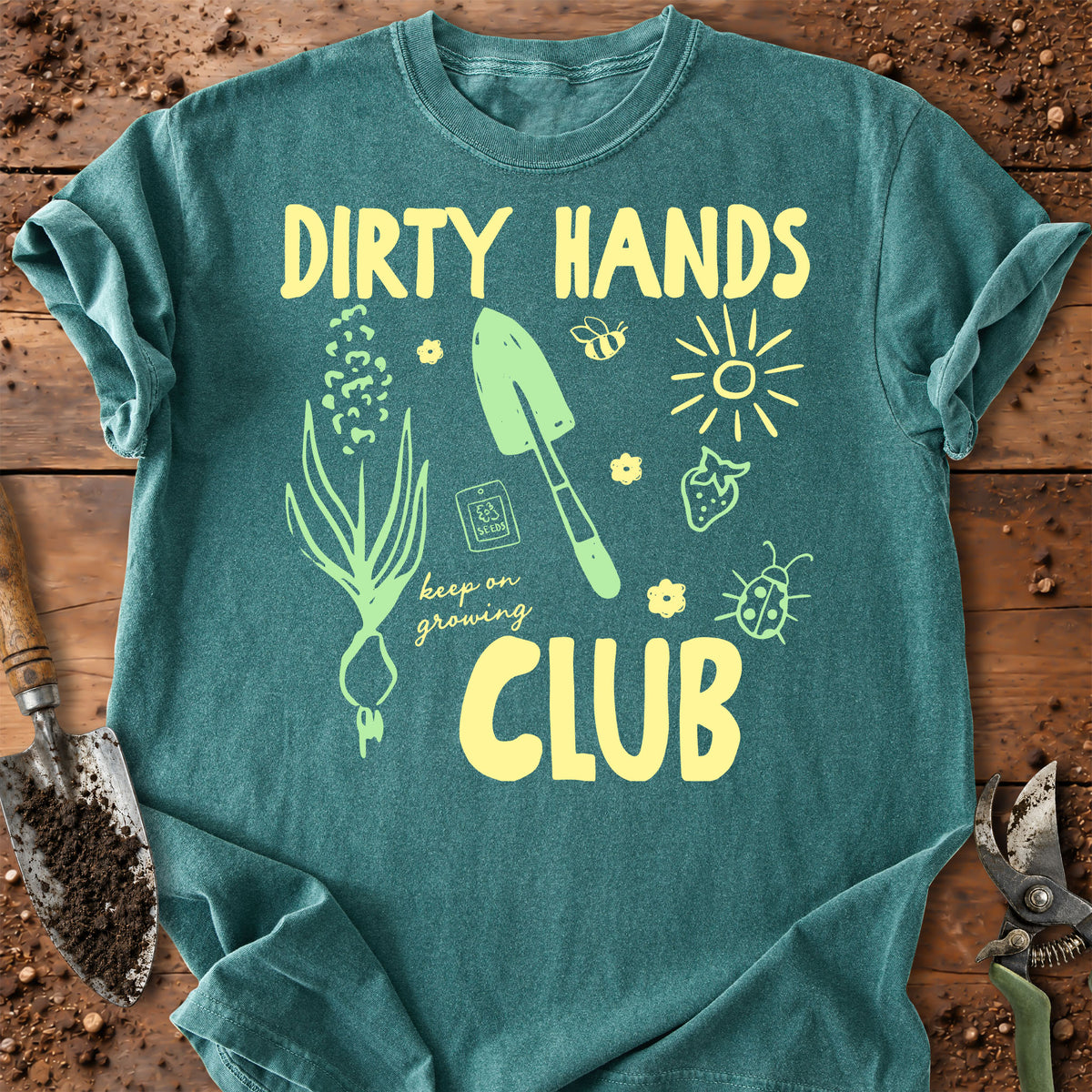 A blue-spruce graphic t-shirt laid flat on a rustic wooden surface with gardening props like soil, a trowel, and pruning shears. The shirt features a yellow and green graphic design with the text 'DIRTY HANDS CLUB' and gardening illustrations.