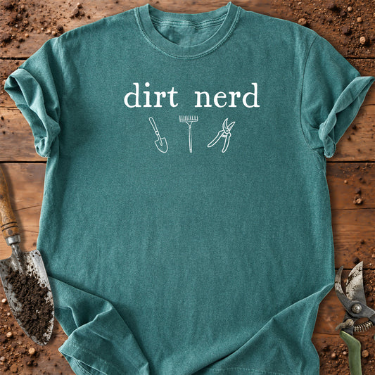 A teal short-sleeve t-shirt with a white graphic that reads 'dirt nerd' above simple line drawings of a shovel, rake, and pruning shears, laid flat on a rustic wooden surface with gardening tools around it.