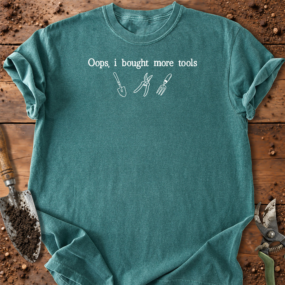 A teal short-sleeve crewneck t-shirt with white text reading 'Oops, i bought more tools' and simple white line drawings of gardening tools, laid flat on a rustic wooden surface with gardening props.