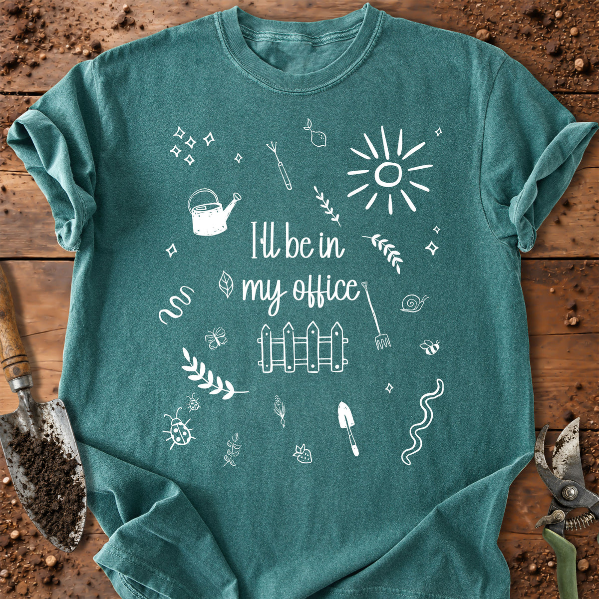 A teal short-sleeve t-shirt with a white graphic print that reads 'I'll be in my office' surrounded by gardening doodles like a watering can, fence, sun, and tools, laid flat on a rustic wooden surface with soil and pruning shears visible.