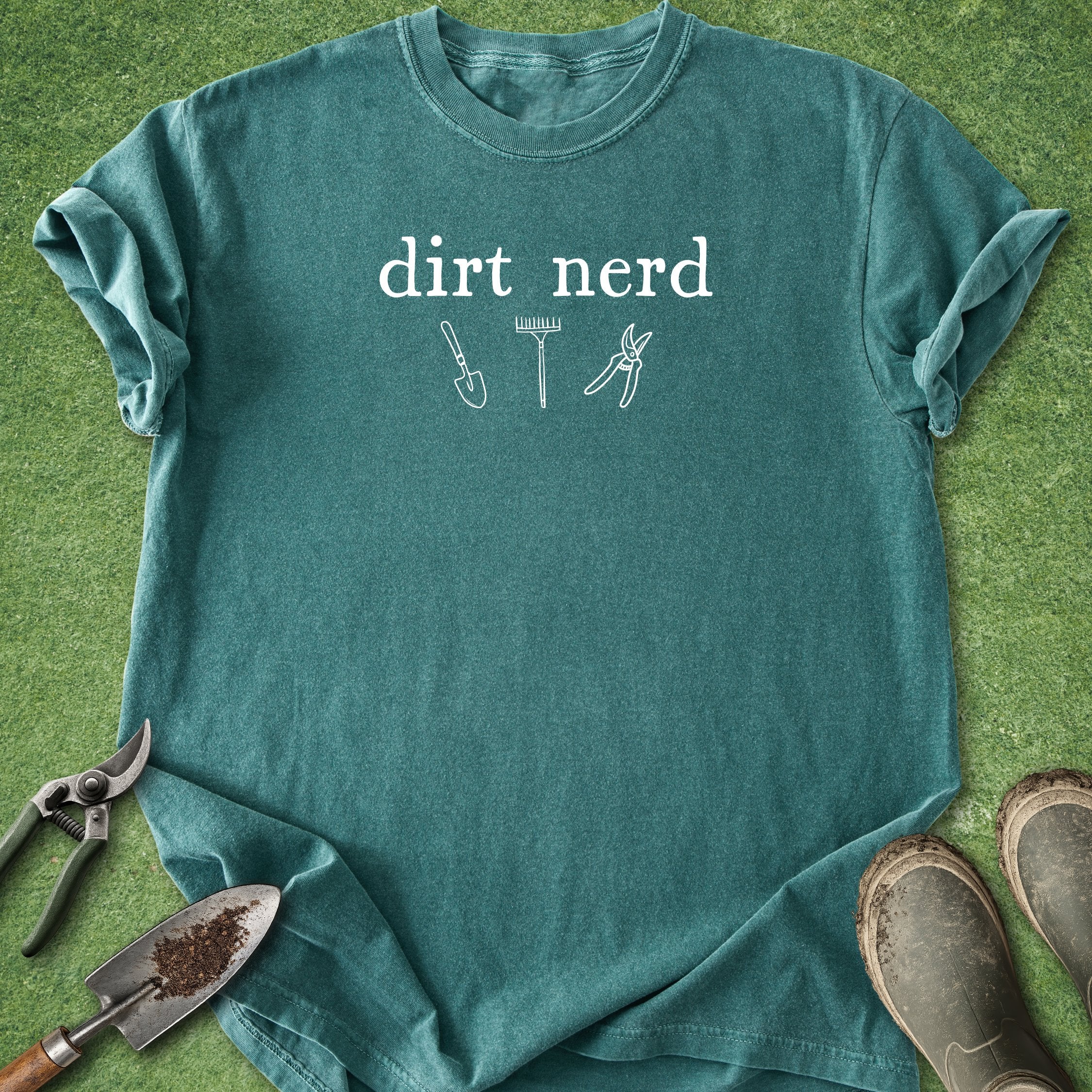Teal t-shirt with 'Oops, I bought more tools' text and gardening tools on a grass background