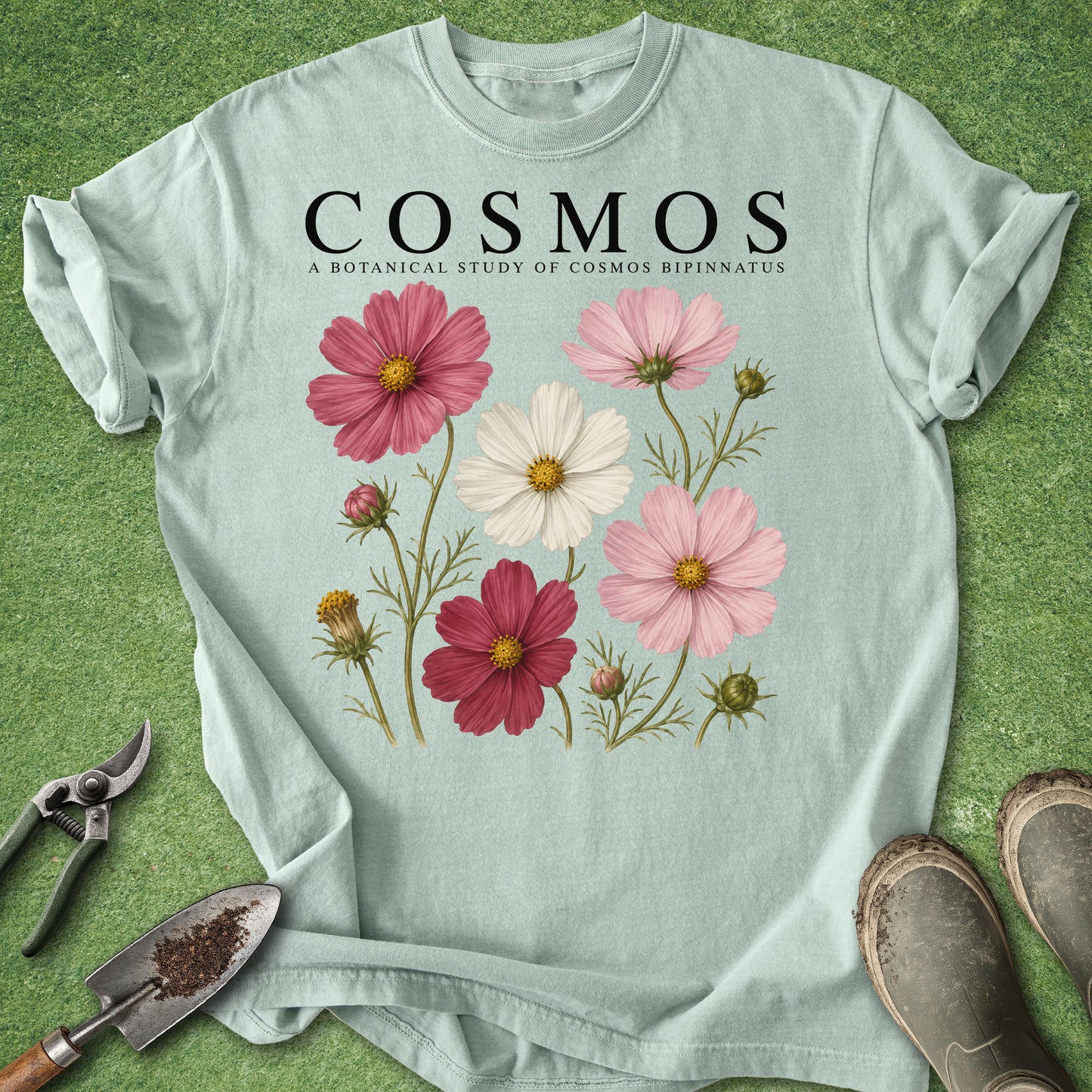 A light mint green graphic t-shirt laid flat on a green grass-like surface, featuring the text 'COSMOS A BOTANICAL STUDY OF COSMOS BIPINNATUS' above a vintage-style floral print with pink, burgundy, and white flowers. A pair of pruning shears, a small trowel, and work boots are placed around the shirt as styling props.
