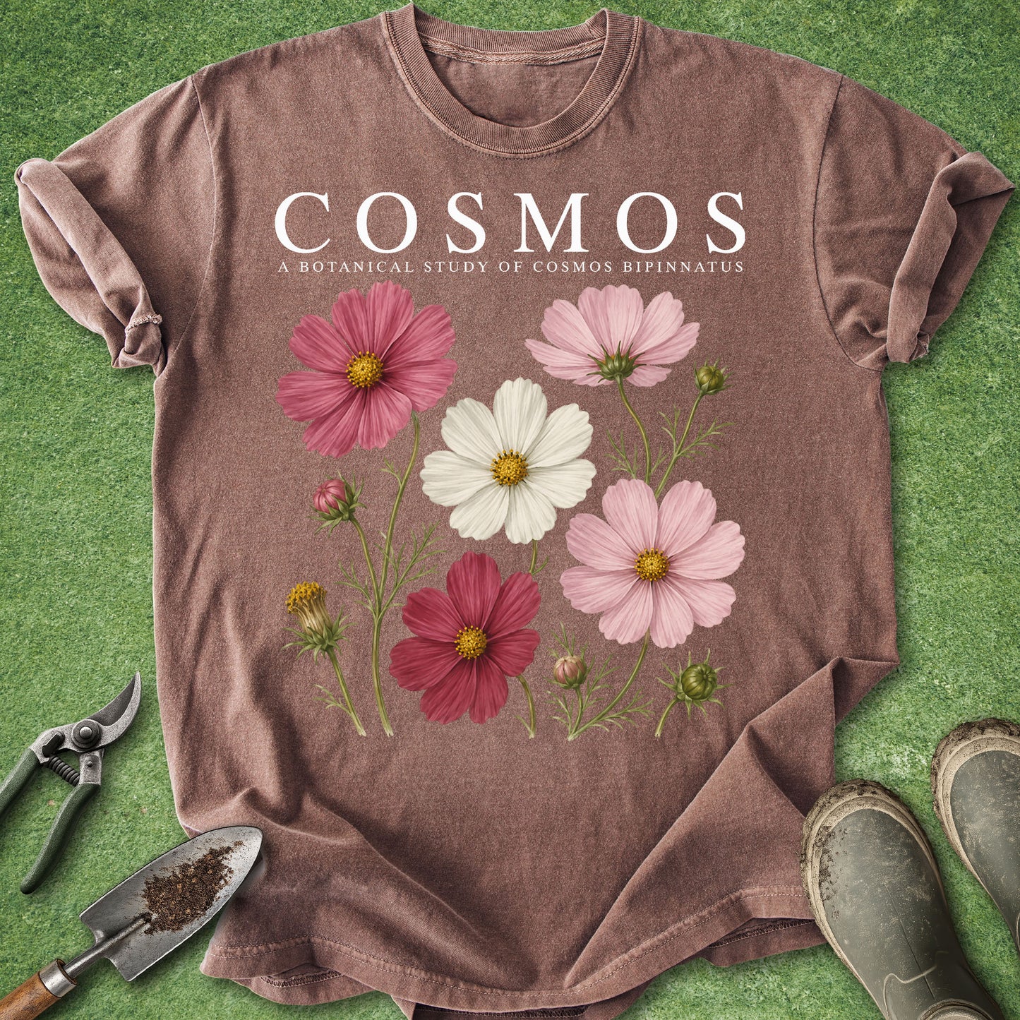 A heathered mauve-brown short-sleeve T-shirt laid flat on green grass-like material, featuring the text 'COSMOS' and smaller botanical text above a cluster of pink, burgundy, and white flowers. Gardening tools and boots are partially visible around the shirt as styled props.
