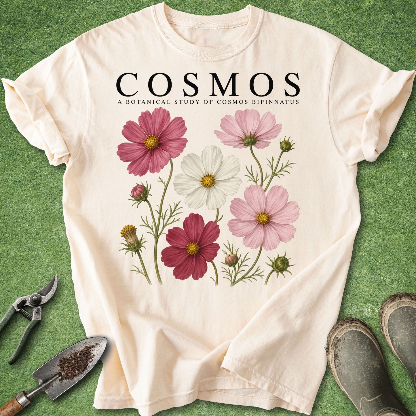 An off-white short-sleeve t-shirt with a botanical graphic of cosmos flowers and the text 'COSMOS A BOTANICAL STUDY OF COSMOS BIPINNATUS' printed on the chest. The shirt is laid flat on a green grass-like surface, surrounded by gardening props including pruning shears, a trowel, and work boots.