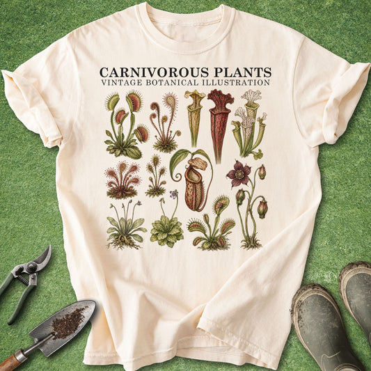 An off-white short-sleeve t-shirt with a vintage botanical illustration of carnivorous plants, laid flat on a green grass-like surface with gardening tools and boots nearby.