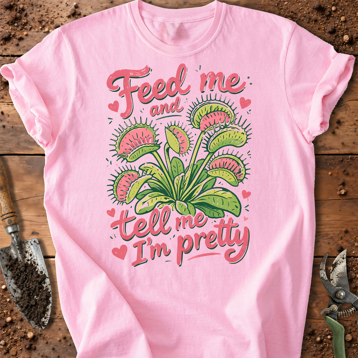 A pink short-sleeve graphic T-shirt laid flat on a rustic wooden surface with gardening props. The shirt features a large illustration of Venus flytraps and the phrase 'Feed me and tell me I'm pretty' in pink lettering with small heart accents.