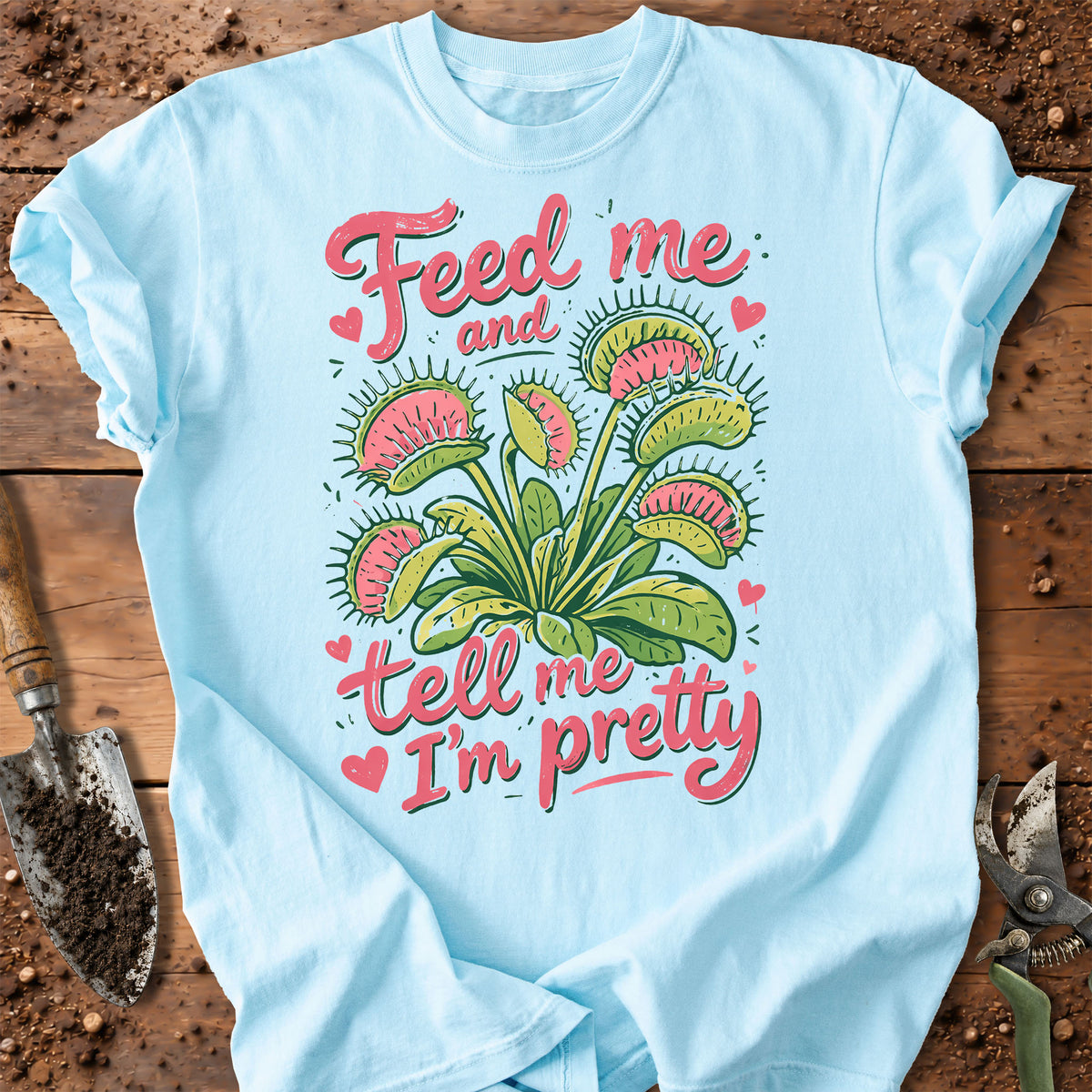 A light blue graphic t-shirt laid flat on a rustic wooden surface with gardening props like a trowel and pruning shears. The shirt features a colorful illustration of a Venus flytrap plant with pink and green details, surrounded by the text 'Feed me and tell me I'm pretty' in pink script with small pink hearts.