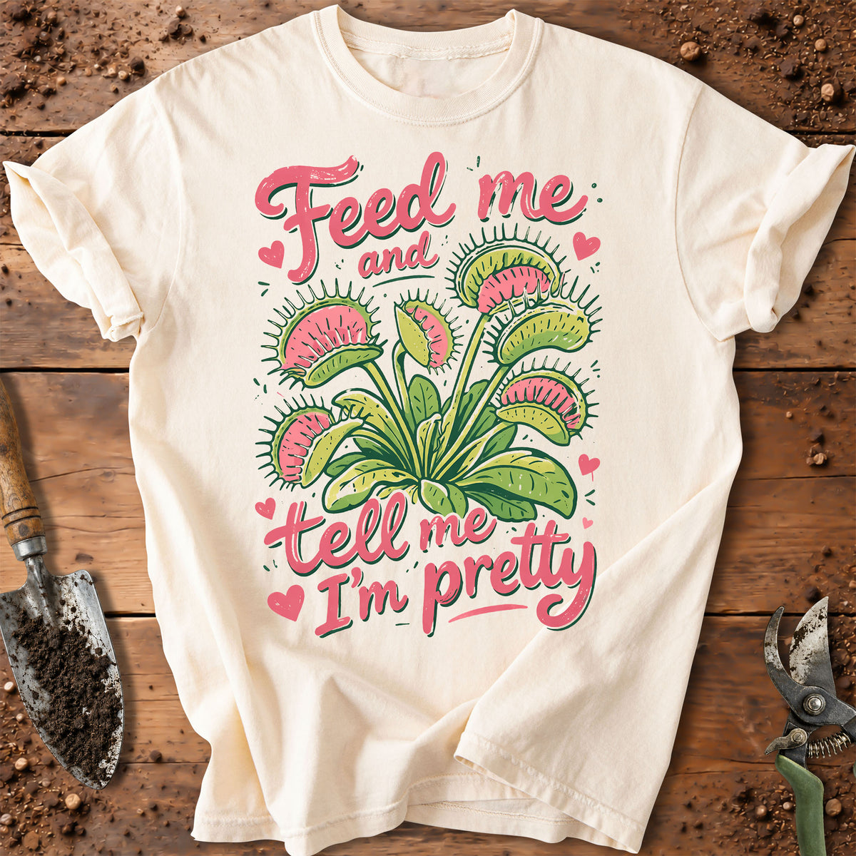 An off-white short-sleeve t-shirt with a graphic of a Venus flytrap plant and the text 'Feed me and tell me I'm pretty' in pink and green, laid flat on a rustic wooden background with gardening tools.