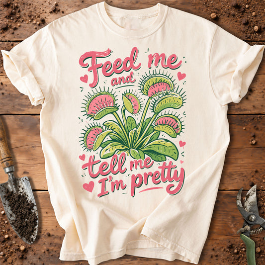 An off-white short-sleeve t-shirt with a graphic of a Venus flytrap plant and the text 'Feed me and tell me I'm pretty' in pink and green, laid flat on a rustic wooden background with gardening tools.