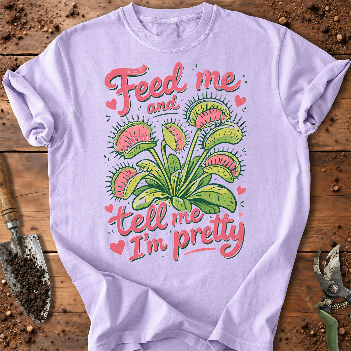 A light lavender crewneck t-shirt with a colorful graphic of a Venus flytrap plant and the text 'Feed me and tell me I'm pretty' in pink, styled in a flat-lay on a wooden surface with gardening tools.