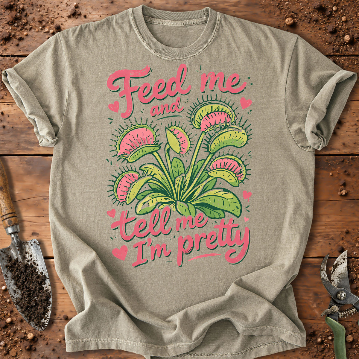 A light beige graphic t-shirt laid flat on a rustic wooden background with gardening tools. The shirt features a colorful illustration of a Venus flytrap plant in green and pink, with the text 'Feed me and tell me I'm pretty' in pink script.