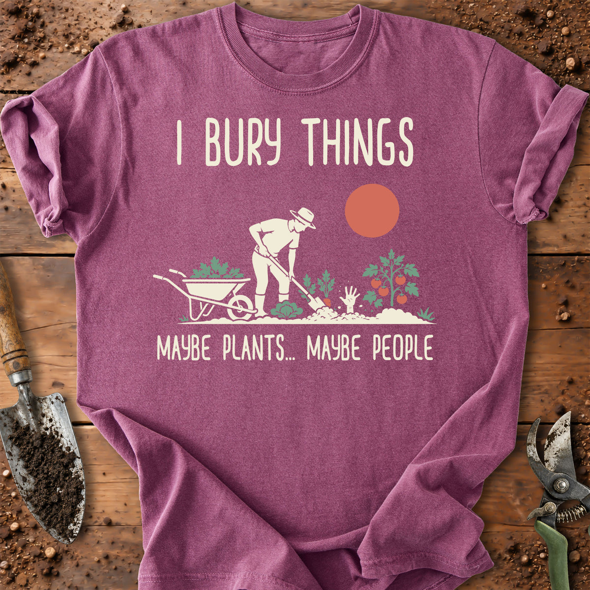 A mauve purple short-sleeve t-shirt laid flat on a rustic wooden surface with gardening props like a trowel, pruning shears, and soil. The shirt features a graphic of a gardener burying a wheelbarrow with the text 'I BURY THINGS' above and 'MAYBE PLANTS.