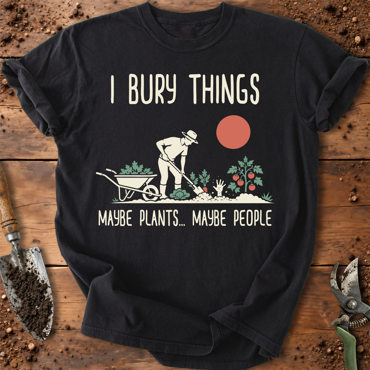 A black graphic t-shirt laid flat on a rustic wooden surface with gardening props like a trowel, soil, and pruning shears. The shirt features a white graphic of a gardener, a wheelbarrow, plants, and a red sun, with the text 'I BURY THINGS' above and 'MAYBE PLANTS.