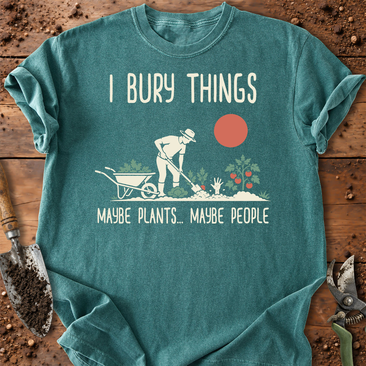 A teal graphic t-shirt laid flat on a rustic wooden surface with gardening props. The shirt features a white illustration of a gardener burying a wheelbarrow and plants, with the text 'I BURY THINGS' above and 'MAYBE PLANTS.