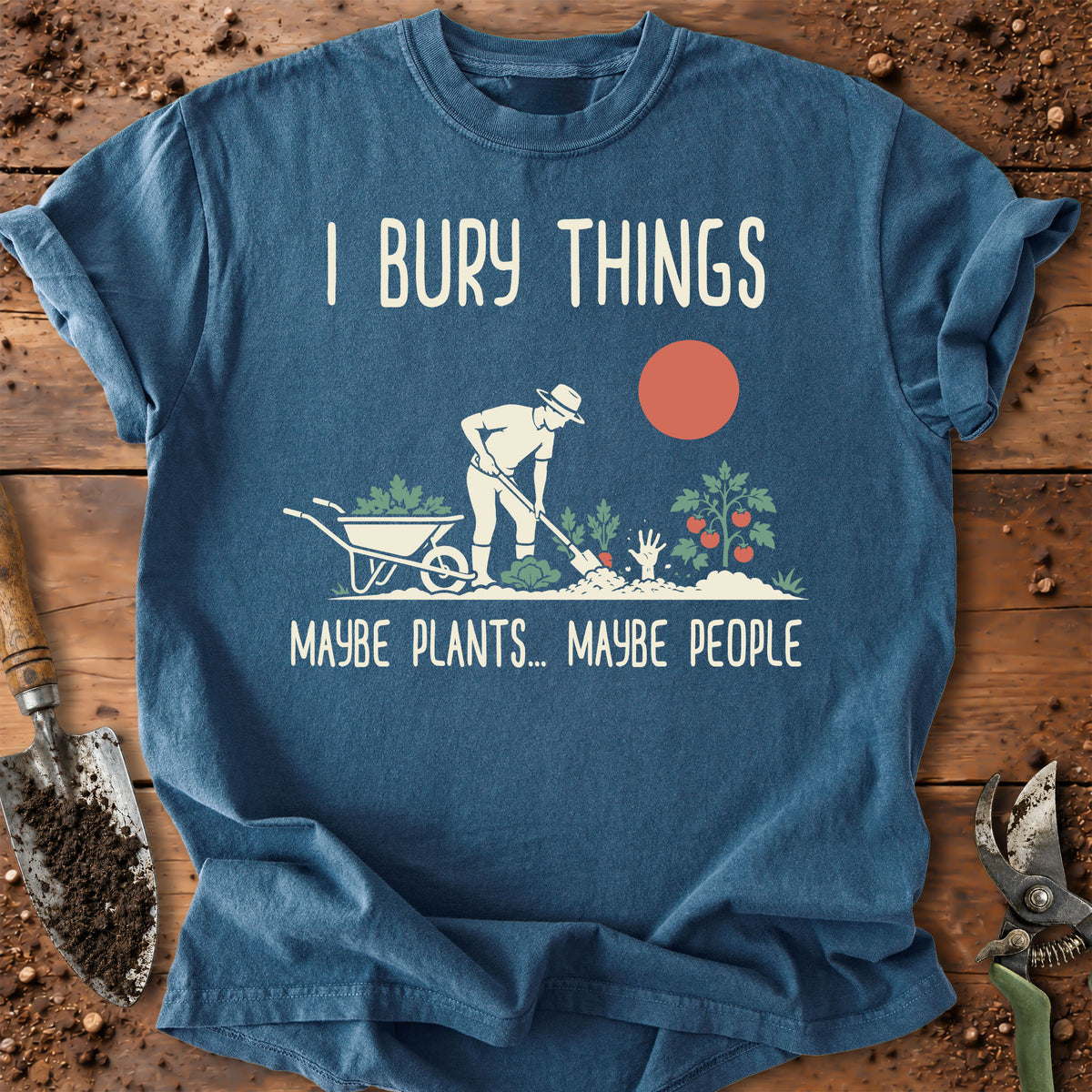 A blue graphic t-shirt laid flat on a rustic wooden surface with gardening props. The shirt features a white graphic of a gardener, plants, and a wheelbarrow, with the text 'I BURY THINGS' above and 'MAYBE PLANTS.