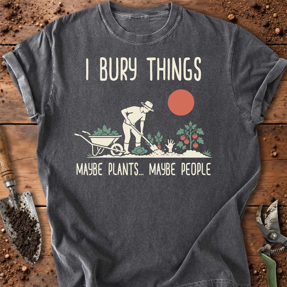 A dark gray graphic t-shirt laid flat on a rustic wooden surface with gardening props. The shirt features a humorous design with the text 'I BURY THINGS' and 'MAYBE PLANTS.