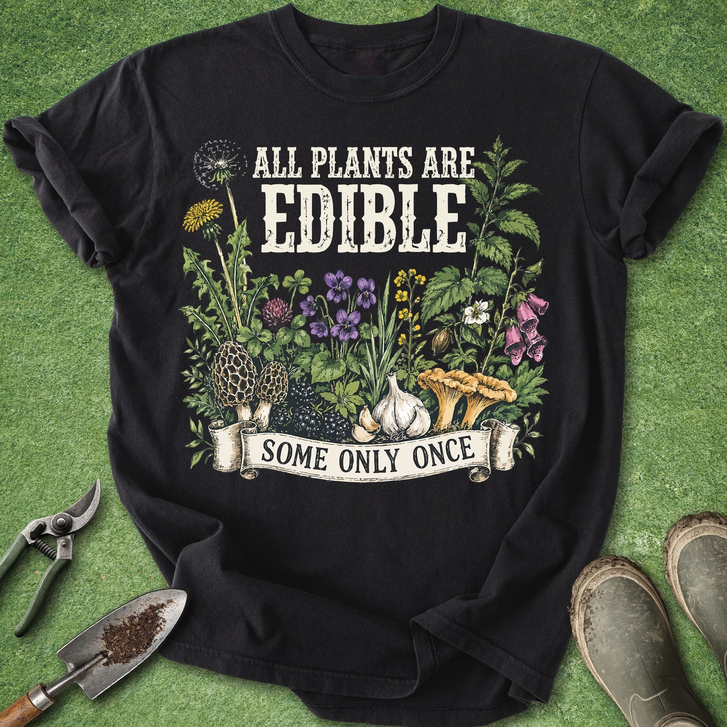 A black graphic t-shirt laid flat on a green grass-like surface, surrounded by gardening props including pruning shears, a trowel, and boots. The shirt features a colorful illustration of edible plants, flowers, garlic, and mushrooms with the text 'ALL PLANTS ARE EDIBLE' and a banner reading 'SOME ONLY ONCE'.