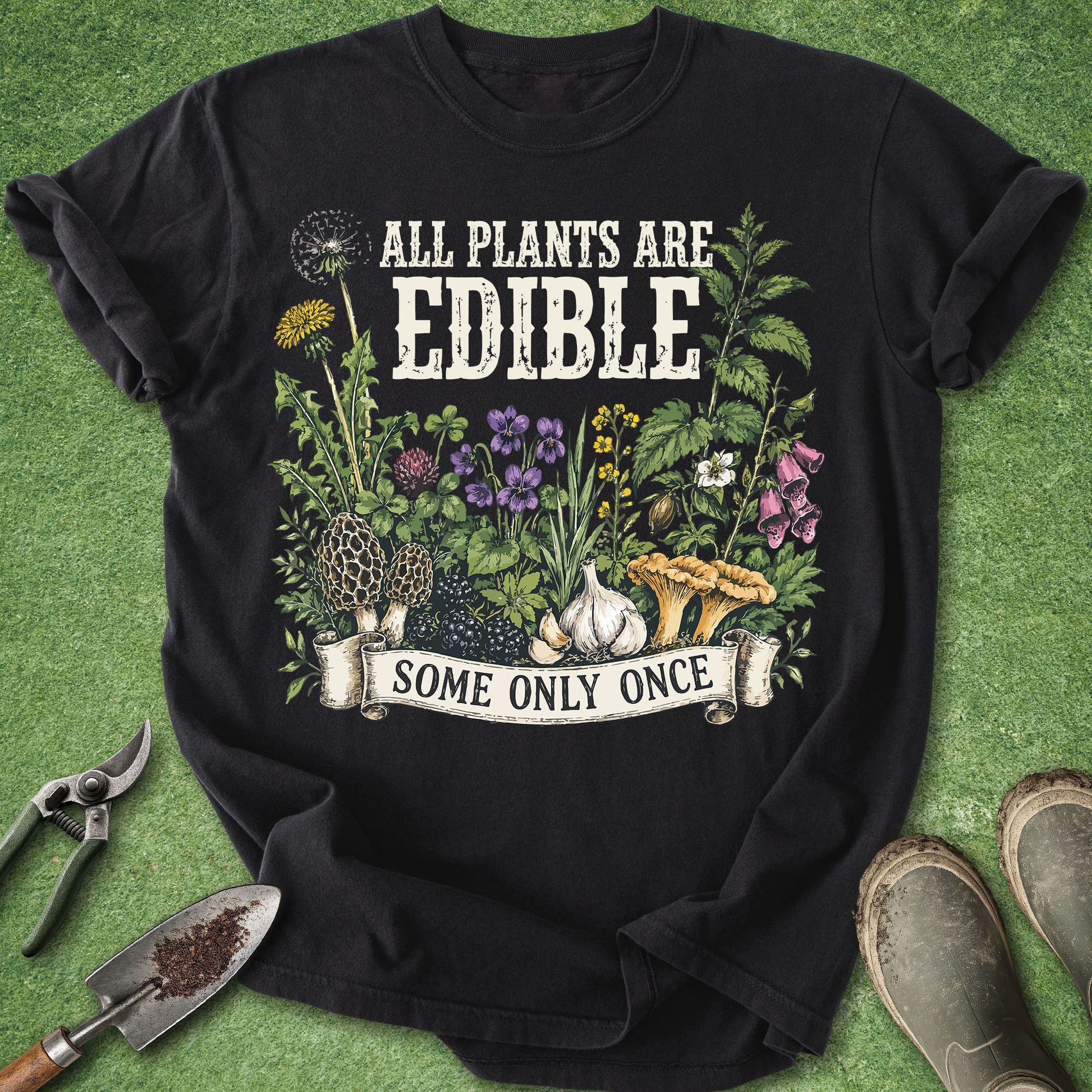 A black graphic t-shirt laid flat on a green grass-like surface, surrounded by gardening props including pruning shears, a trowel, and boots. The shirt features a colorful illustration of edible plants, flowers, garlic, and mushrooms with the text 'ALL PLANTS ARE EDIBLE' and a banner reading 'SOME ONLY ONCE'.