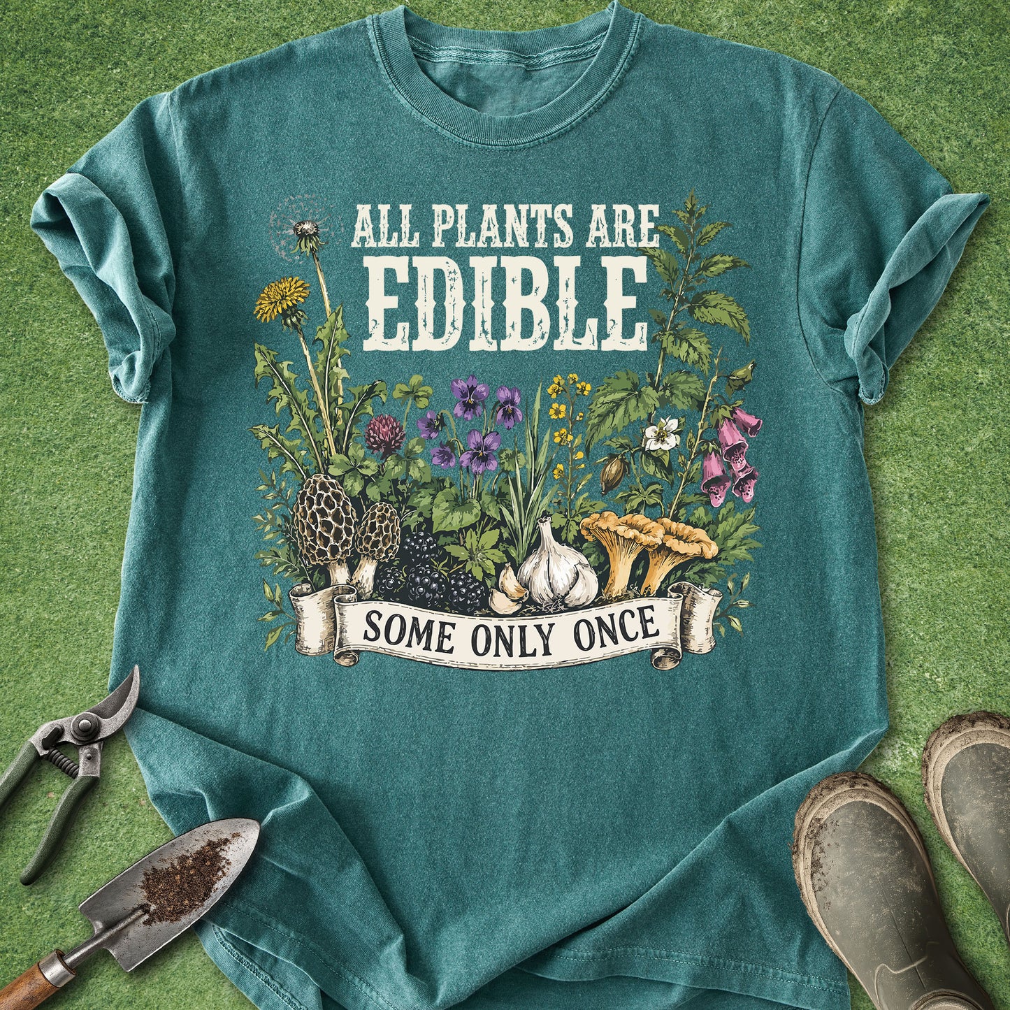 A teal graphic t-shirt laid flat on a green grass-like surface, with gardening props around it: pruning shears, a trowel, and boots. The shirt reads 'ALL PLANTS ARE EDIBLE SOME ONLY ONCE' above a botanical illustration with flowers, mushrooms, garlic, and herbs.