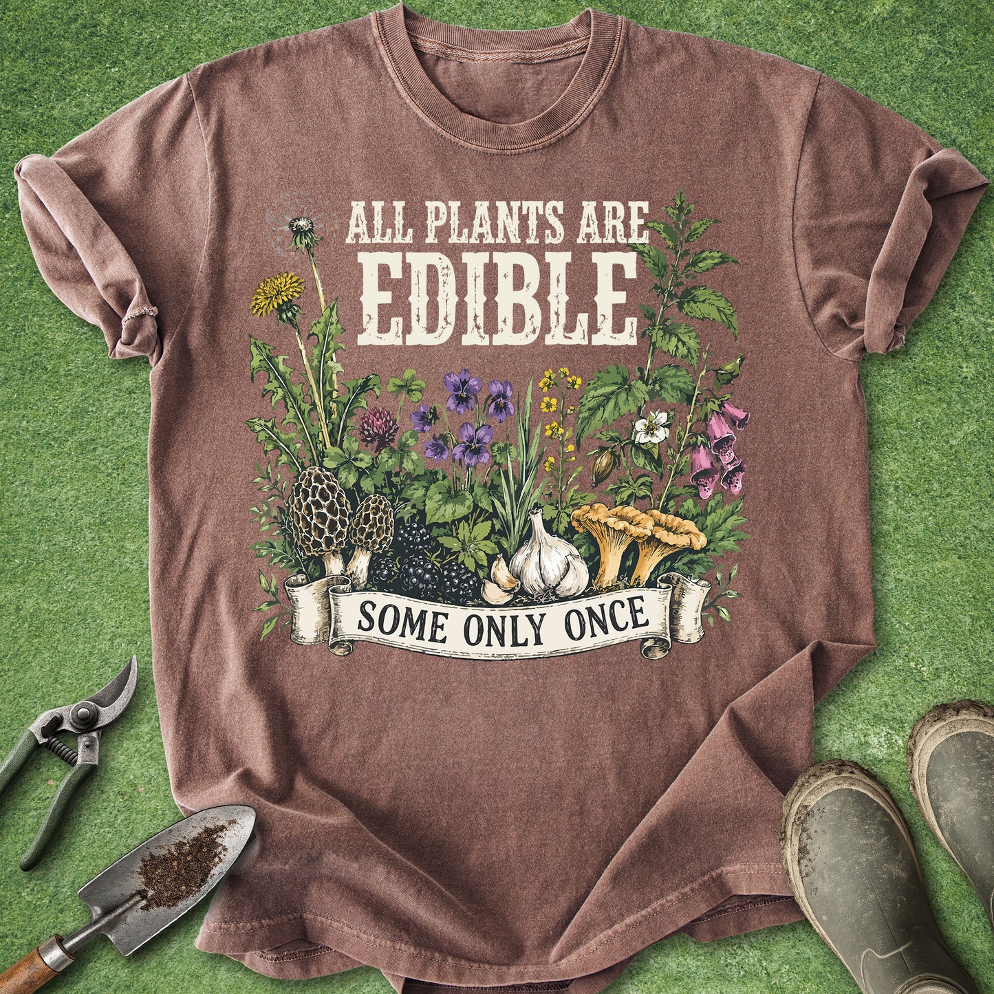 A brown short-sleeve graphic T-shirt laid flat on green grass-like ground, with gardening tools and boots around it. The shirt reads 'ALL PLANTS ARE EDIBLE' above an illustrated garden scene with flowers, mushrooms, garlic, and a banner that says 'SOME ONLY ONCE'.