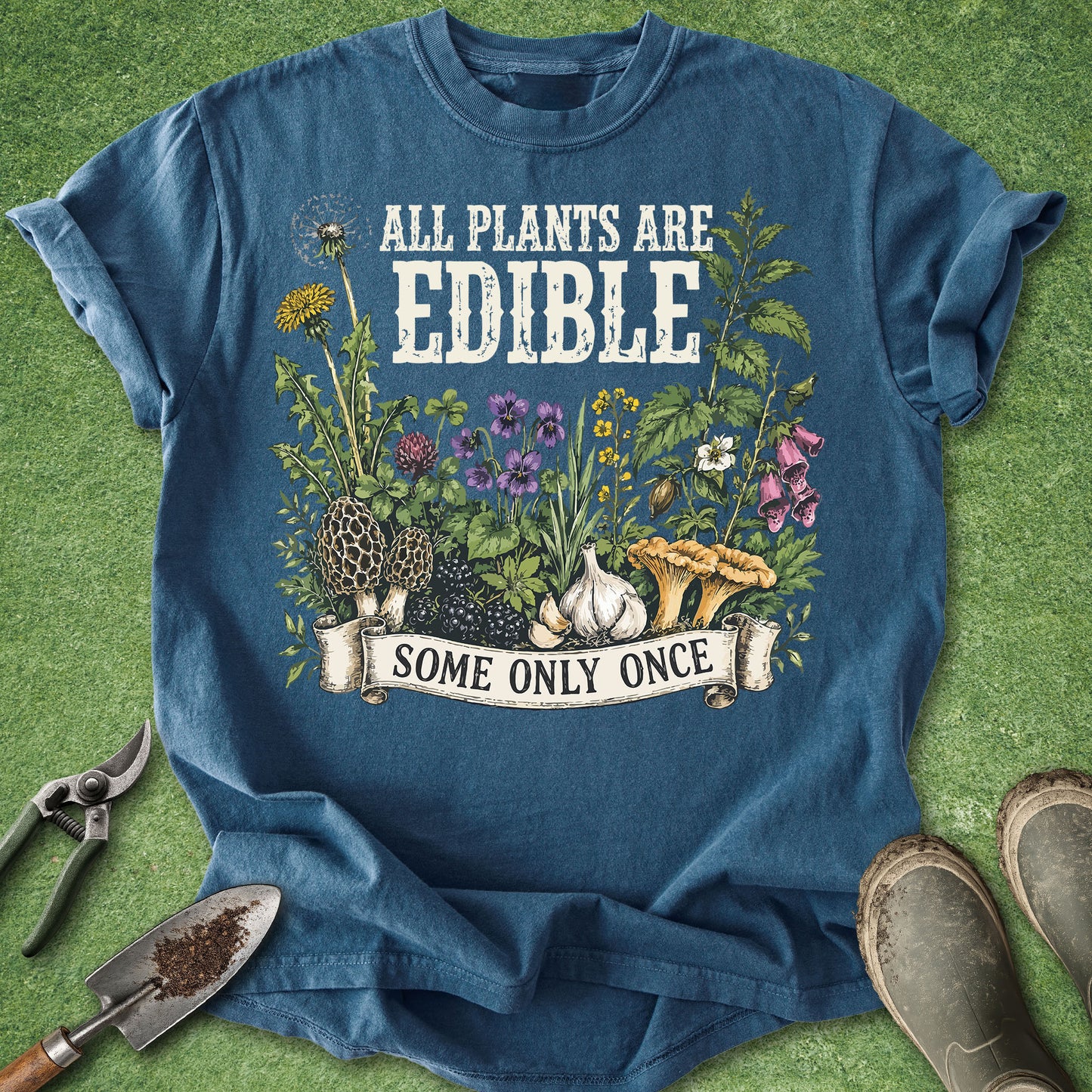 A blue graphic t-shirt laid flat on green grass with gardening props like pruning shears, a trowel, and boots. The shirt features a white text graphic reading 'ALL PLANTS ARE EDIBLE SOME ONLY ONCE' above an illustration of edible plants, flowers, garlic, and mushrooms.