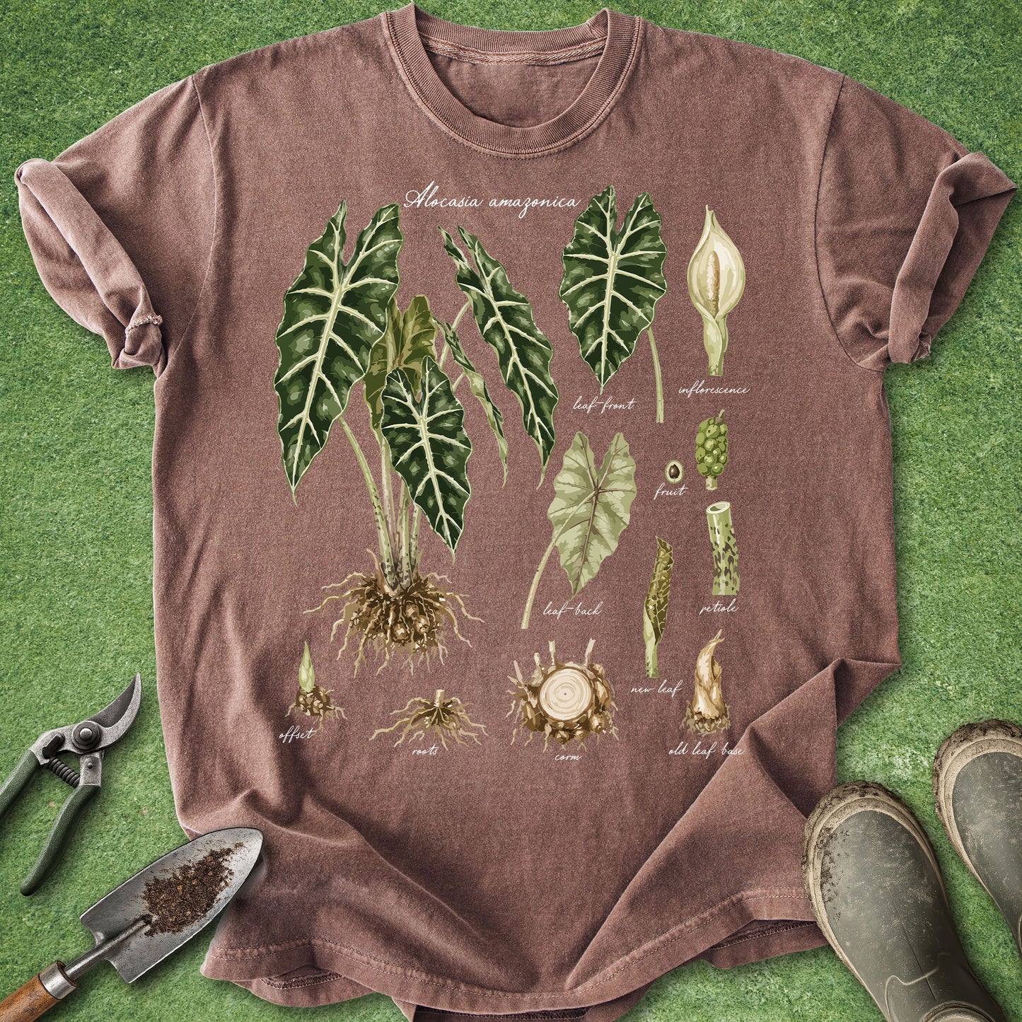 A heathered brown short-sleeve t-shirt laid flat on green grass, featuring a botanical illustration of Alocasia amazonica plant parts. Gardening props including pruning shears, a trowel, and boots are visible around the shirt.