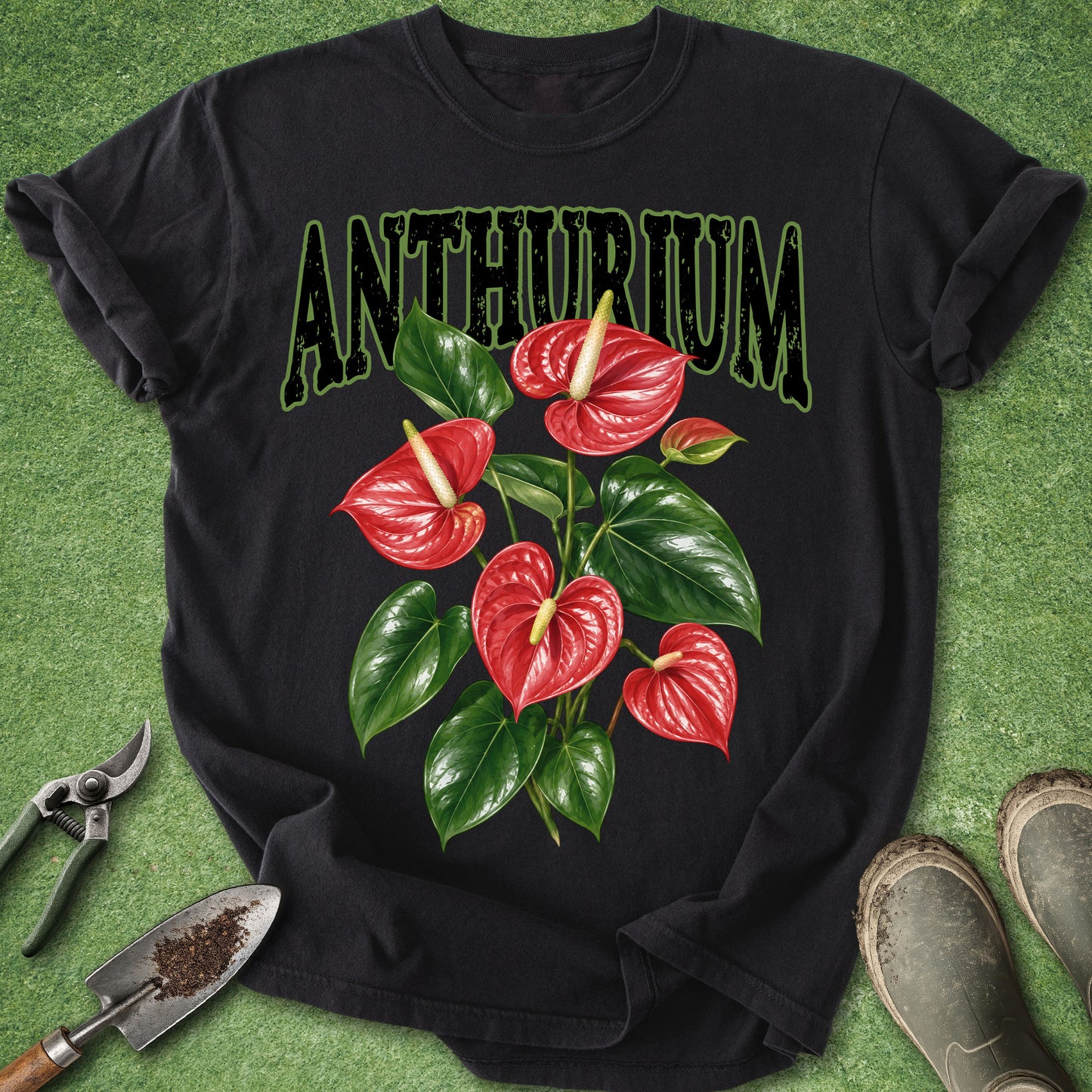A black short-sleeve graphic T-shirt laid flat on green artificial turf, featuring the word 'ANTHURIUM' above a cluster of red anthurium flowers and green leaves. Gardening props including pruning shears, a trowel, and boots are placed around the shirt.
