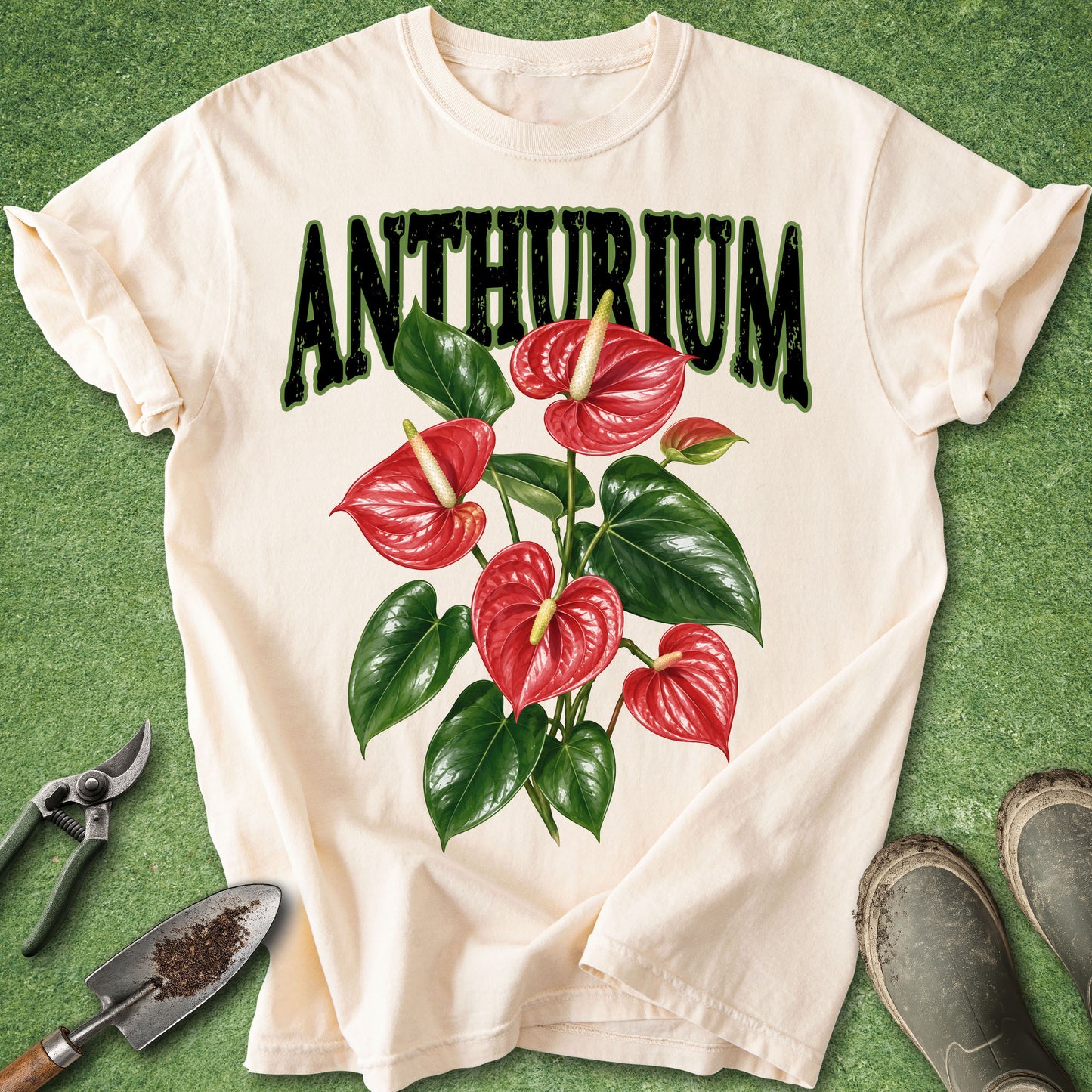 An off-white short-sleeve t-shirt laid flat on a green grass-like surface, featuring a large graphic print of red anthurium flowers and the word 'ANTHURIUM' in dark green letters. Gardening props including pruning shears, a small trowel, and boots are placed around the shirt.