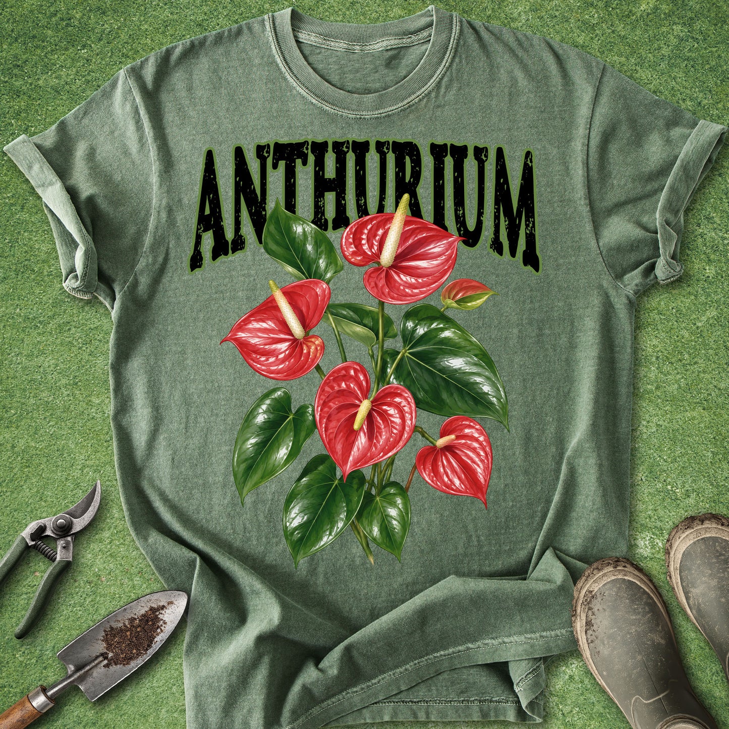 An olive green graphic t-shirt laid flat on a green grass-like surface, featuring the word 'ANTHURIUM' in black distressed letters above a large illustration of red anthurium flowers and green leaves. Gardening tools and boots are partially visible around the shirt as styling props.