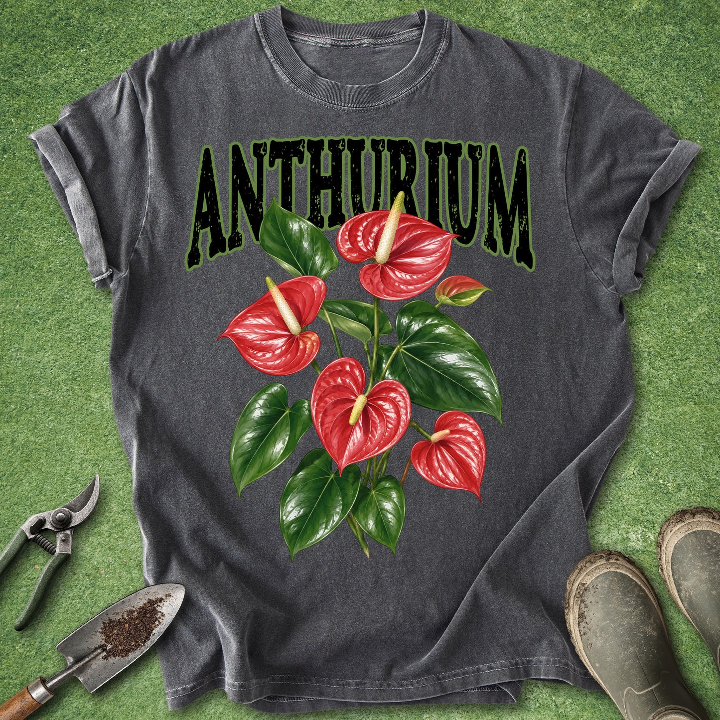 A dark gray t-shirt laid flat on a green grass-like surface, featuring a large graphic of red anthurium flowers and green leaves with the word 'ANTHURIUM' in black outlined letters. Gardening tools and boots are placed around the shirt as styling props.