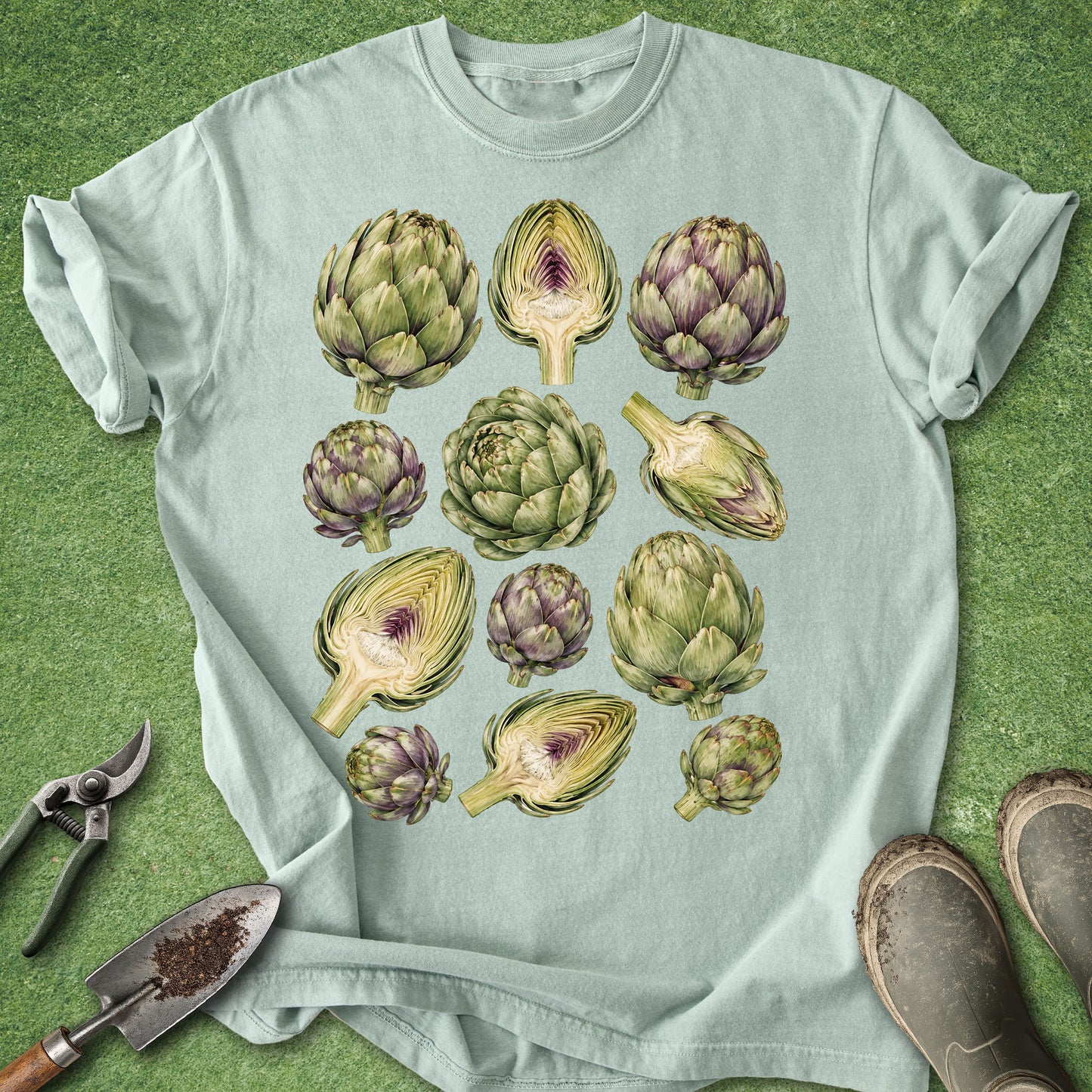 A light sage green t-shirt with a graphic print of various artichokes, laid flat on a green grass-like surface with gardening props like pruning shears, a trowel, and boots.