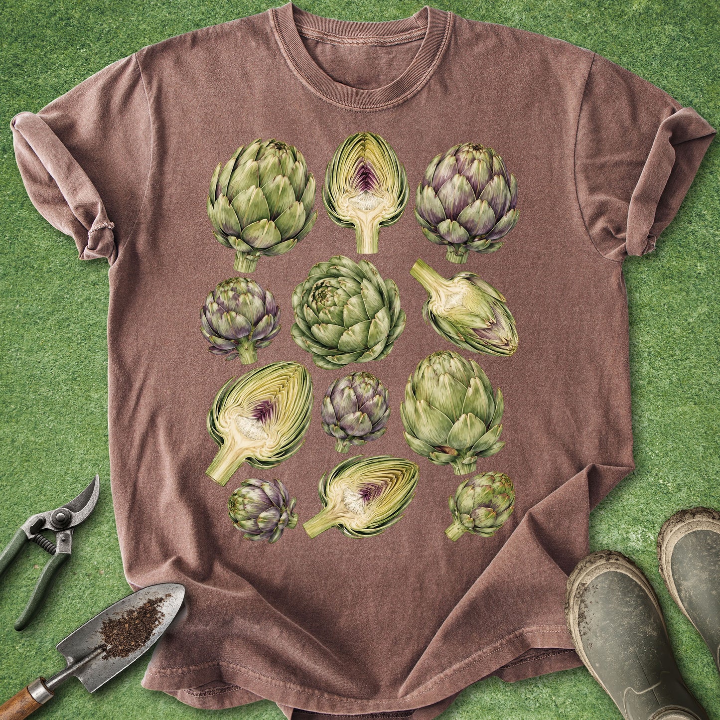 A brown short-sleeve t-shirt with a graphic print of various green artichokes, laid flat on a green grass-like surface with gardening tools and boots nearby.