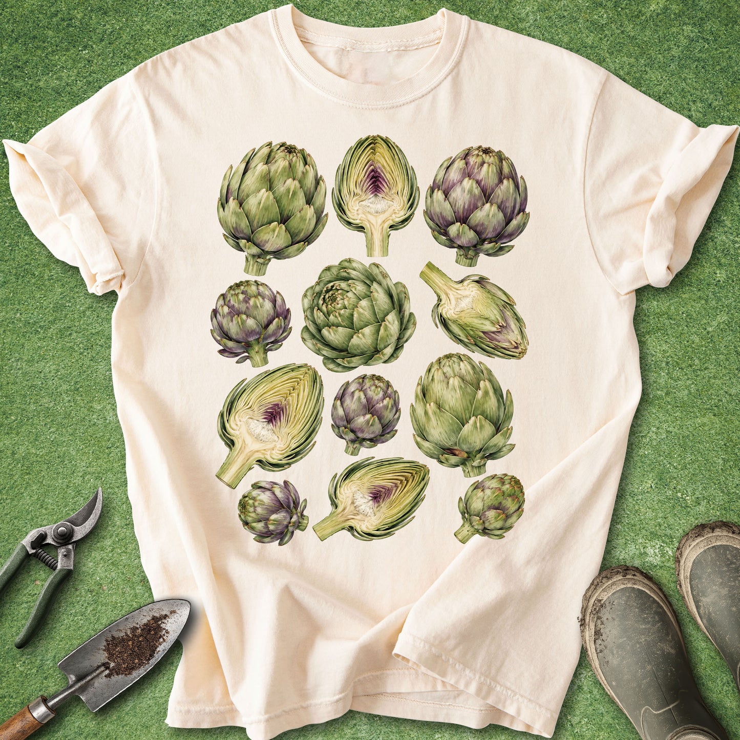 A cream-colored short-sleeve t-shirt with a graphic print of various green and purple artichokes, laid flat on a green grass-like background with gardening tools and boots nearby.