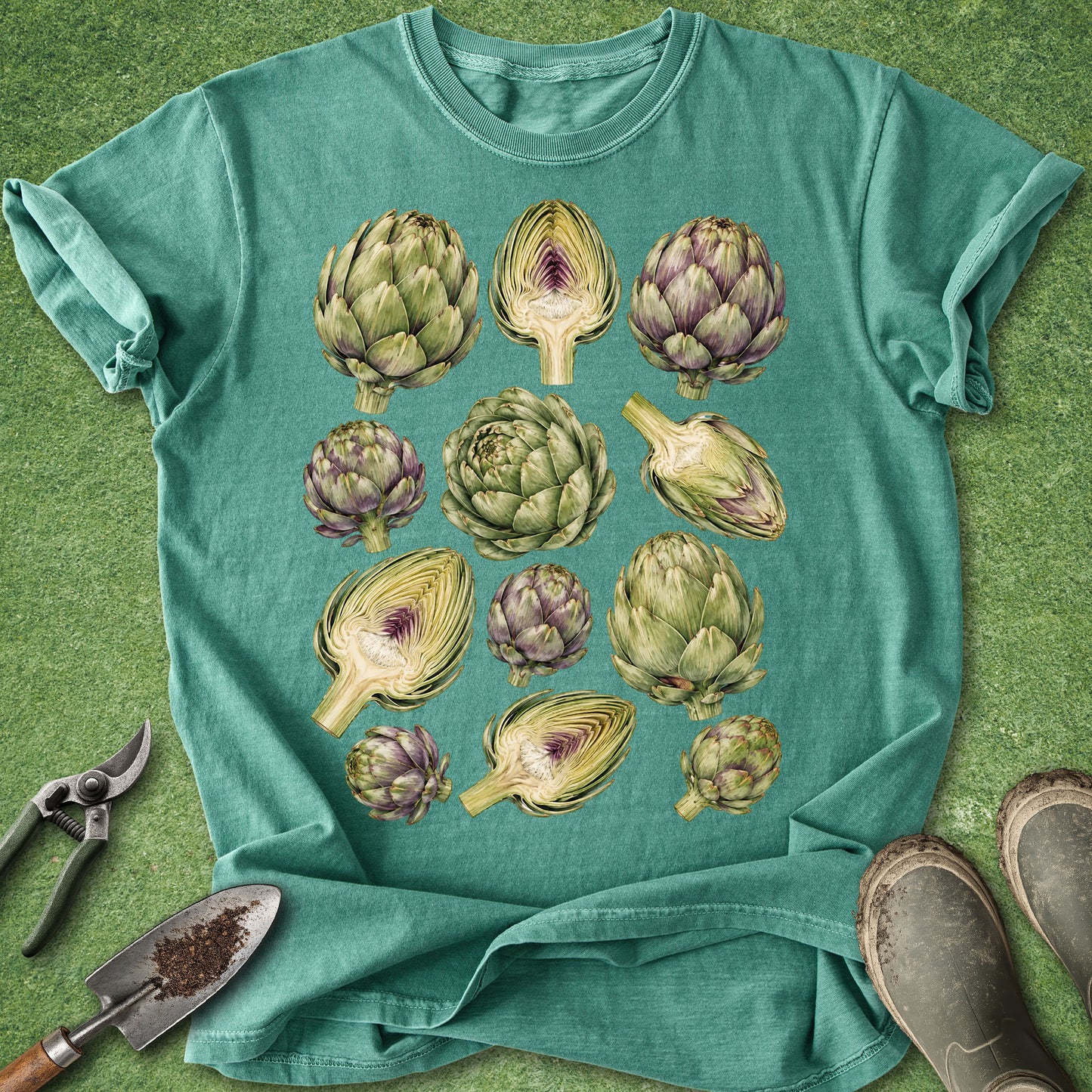 A teal short-sleeve t-shirt laid flat on a green grass-like surface, featuring a large graphic print of multiple artichokes in green and purple tones. Gardening props including pruning shears, a trowel, and boots are placed around the shirt.