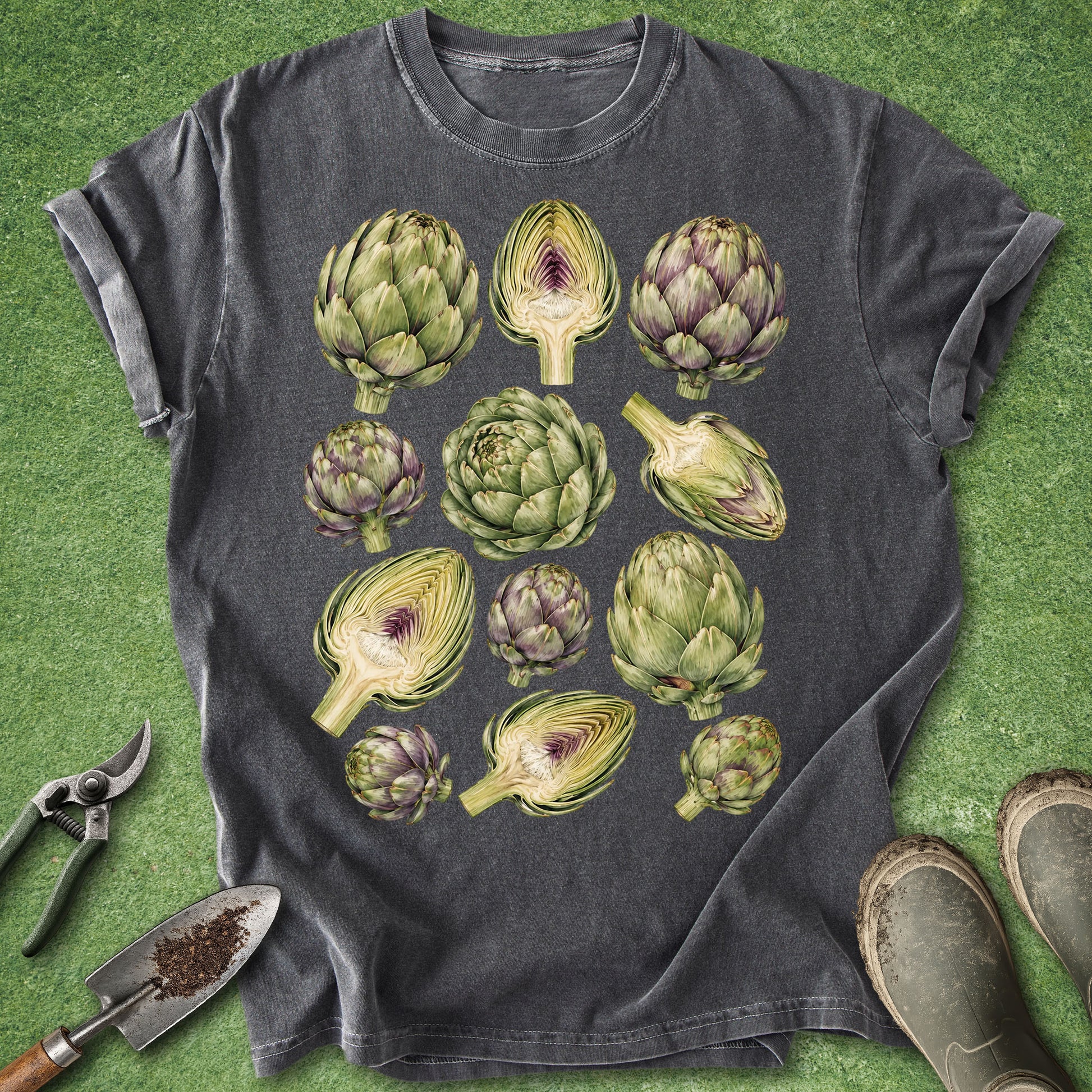 A dark gray t-shirt with a graphic print of various green and purple artichokes, laid flat on a green grass-like background with gardening tools and boots around it.