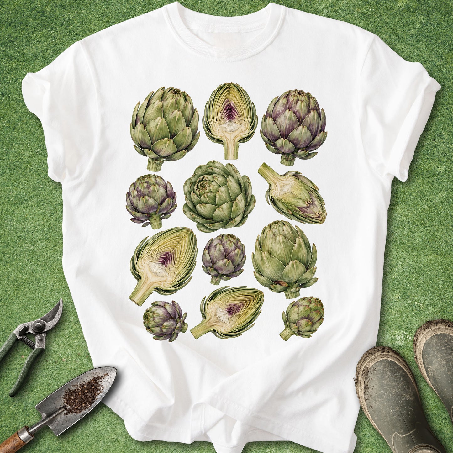 A white short-sleeve T-shirt laid flat on a green grass-like surface, featuring a large printed collage of illustrated artichokes in green and purple tones. Gardening props, including pruning shears, a small hand trowel, and muddy boots, are placed around the shirt.