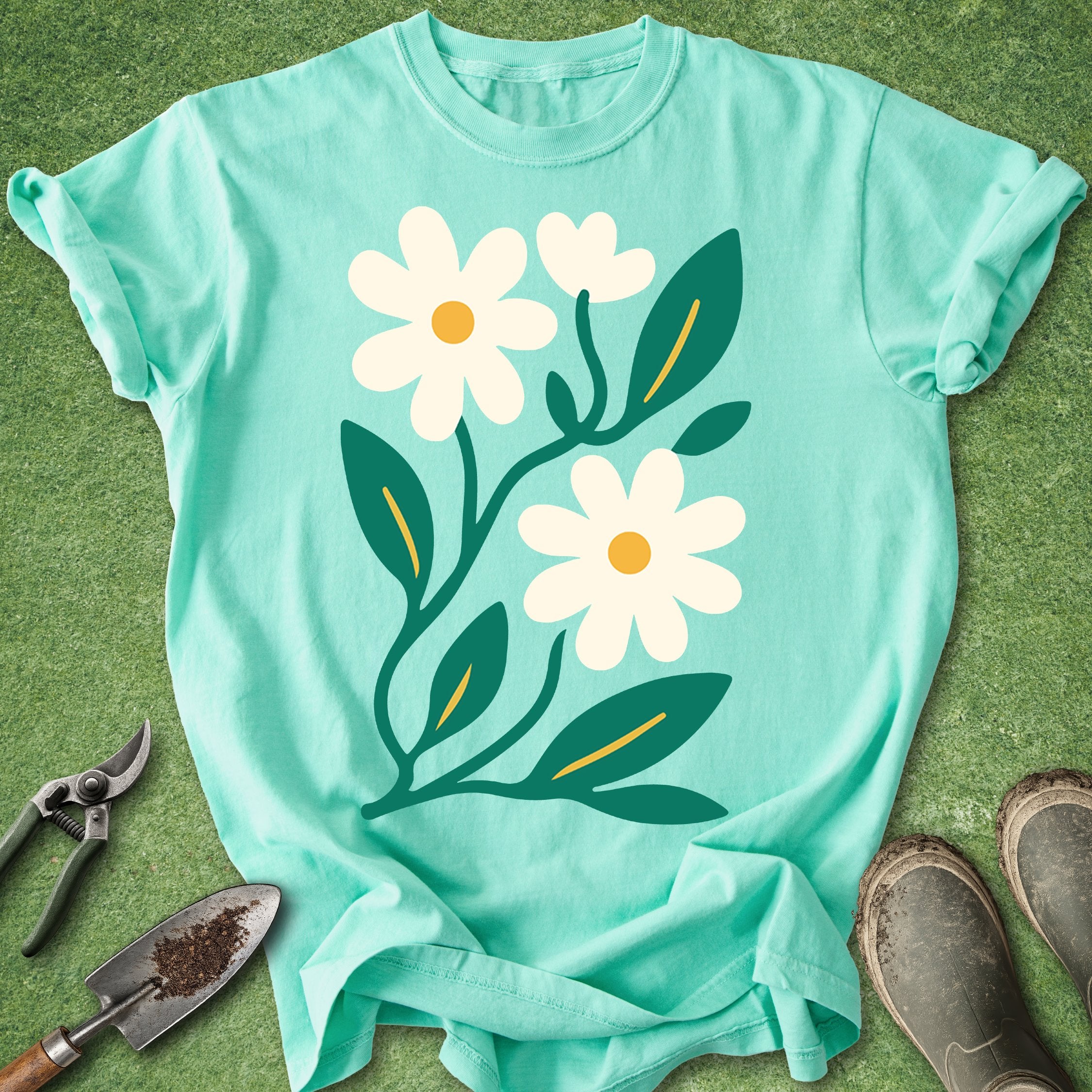 Teal t-shirt with floral design on a green surface with gardening tools.