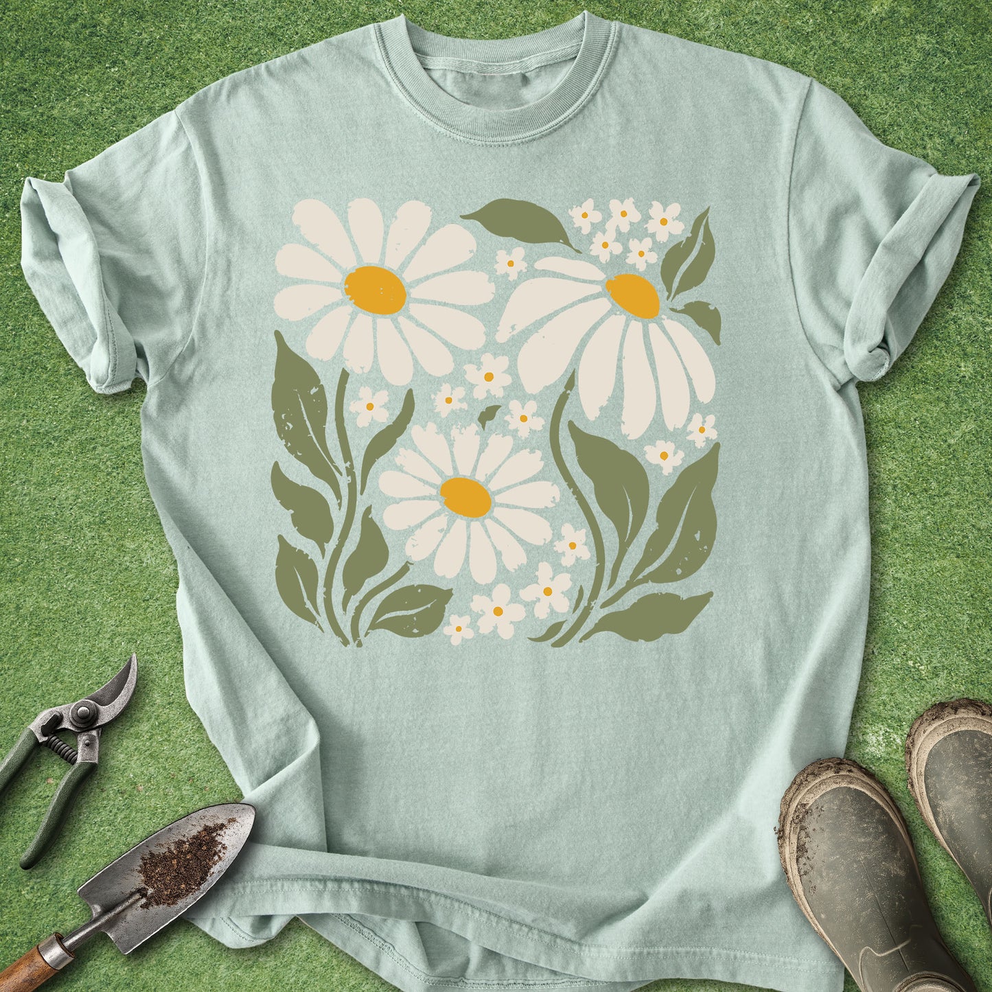 A light sage green t-shirt with a retro-style floral print of white daisies and green leaves, laid flat on a green grass background with gardening tools and boots nearby.