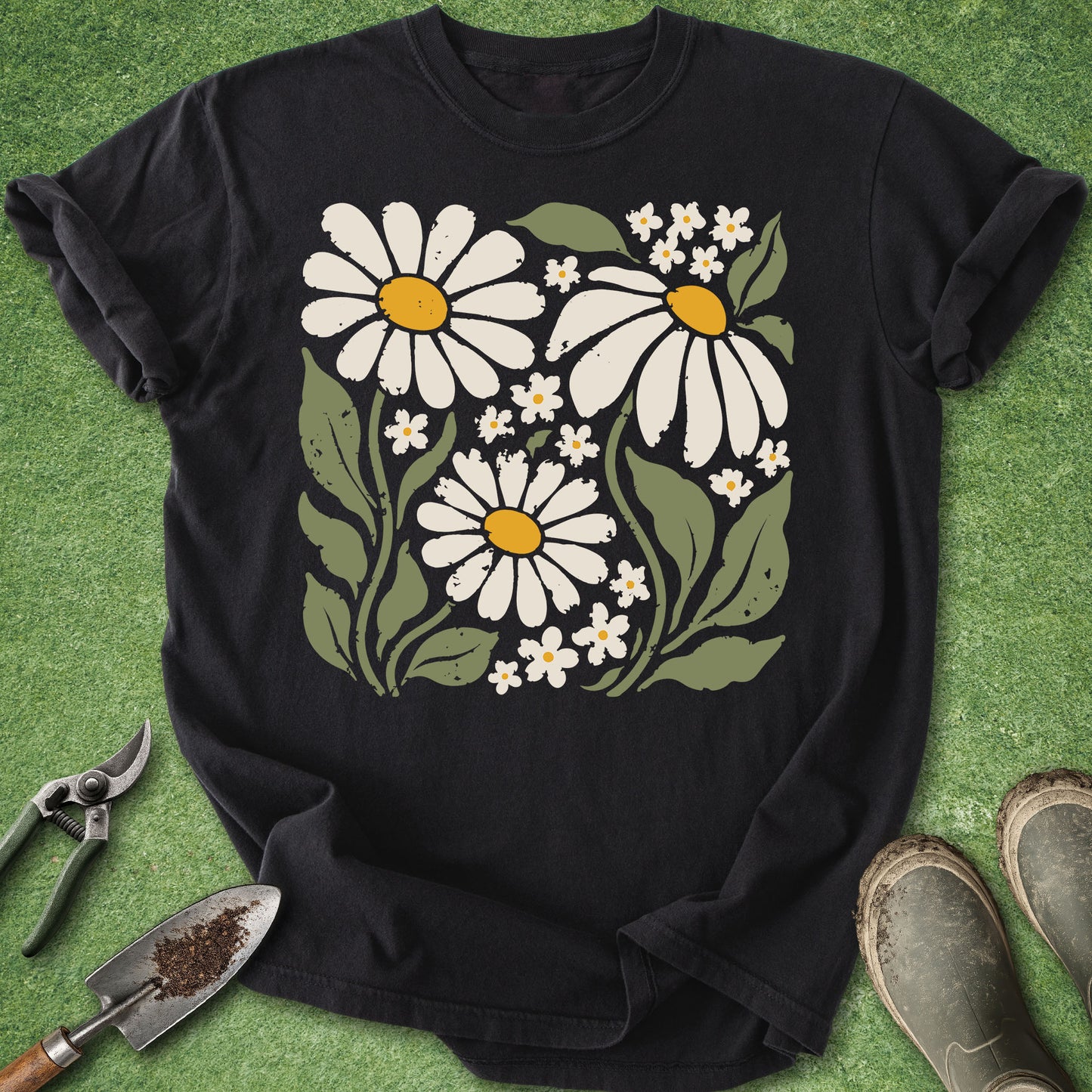 A black short-sleeve t-shirt laid flat on green grass, featuring a large graphic print of white daisies with yellow centers and green leaves. Gardening tools and muddy boots are placed around the shirt as props.