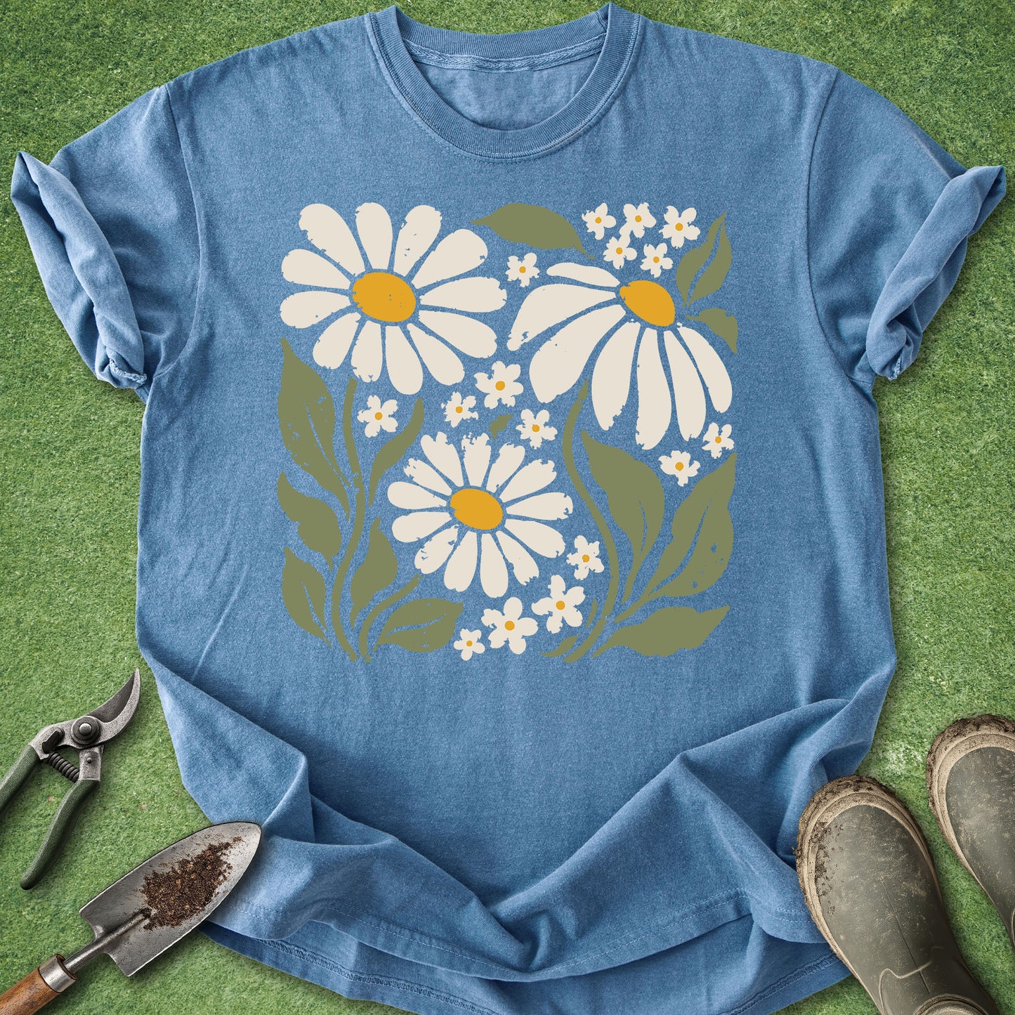 A blue short-sleeve crewneck t-shirt with a retro-style daisy and flower graphic, laid flat on green grass with gardening tools and boots nearby.