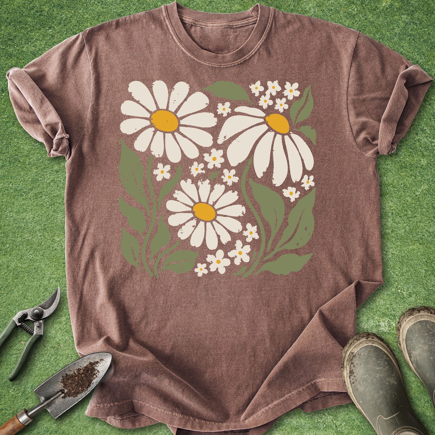 A brown short-sleeve t-shirt with a daisy and wildflower graphic, laid flat on green artificial grass with gardening tools and boots as props.
