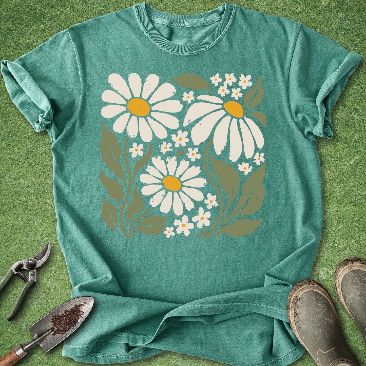 A teal short-sleeve t-shirt with a graphic of white daisies with yellow centers and green leaves, laid flat on a green grass-like background with gardening tools and boots nearby.